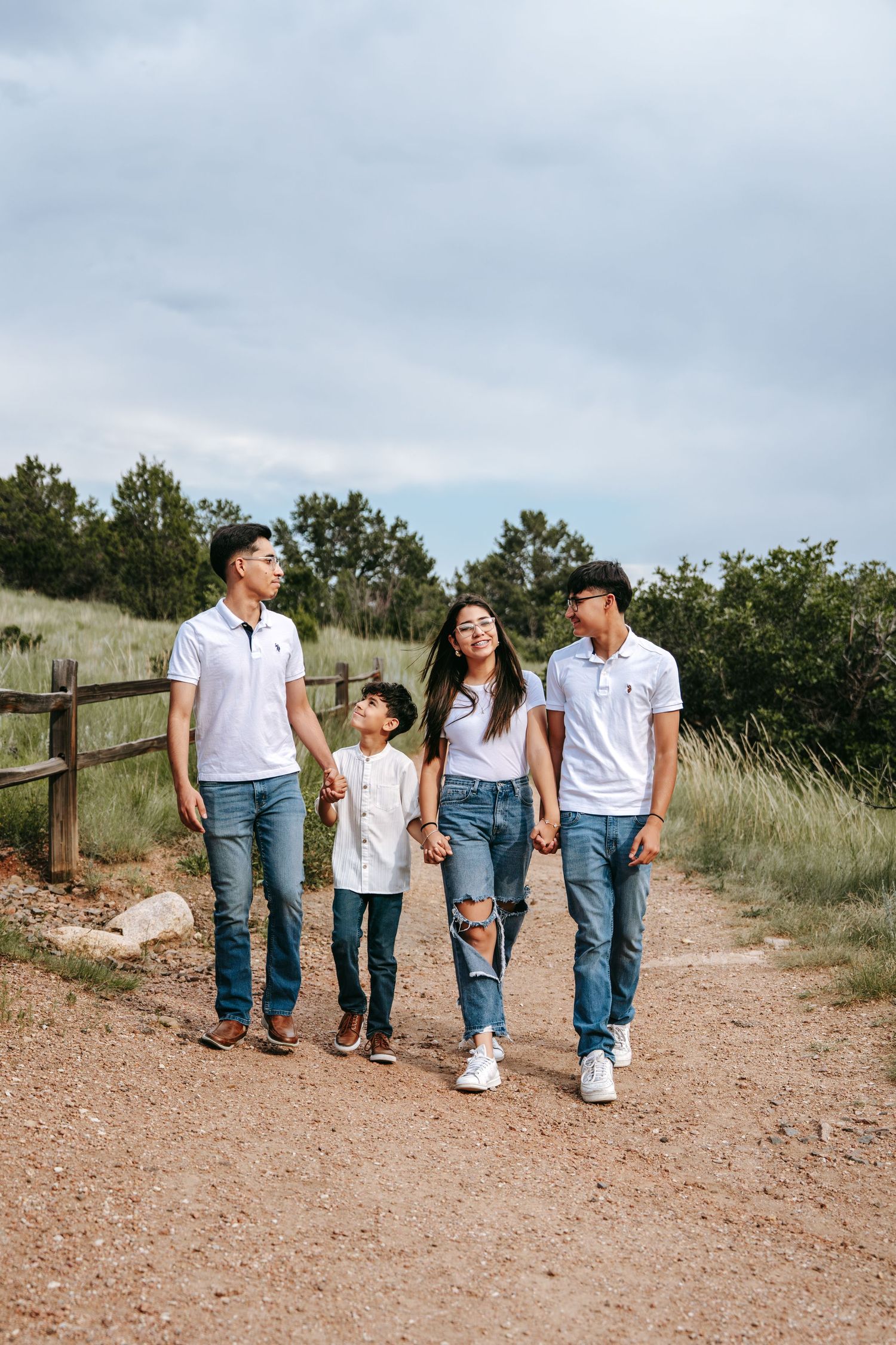 Family of four in white polo shirts and jeans walking together on a dirt trail with green fields and trees.