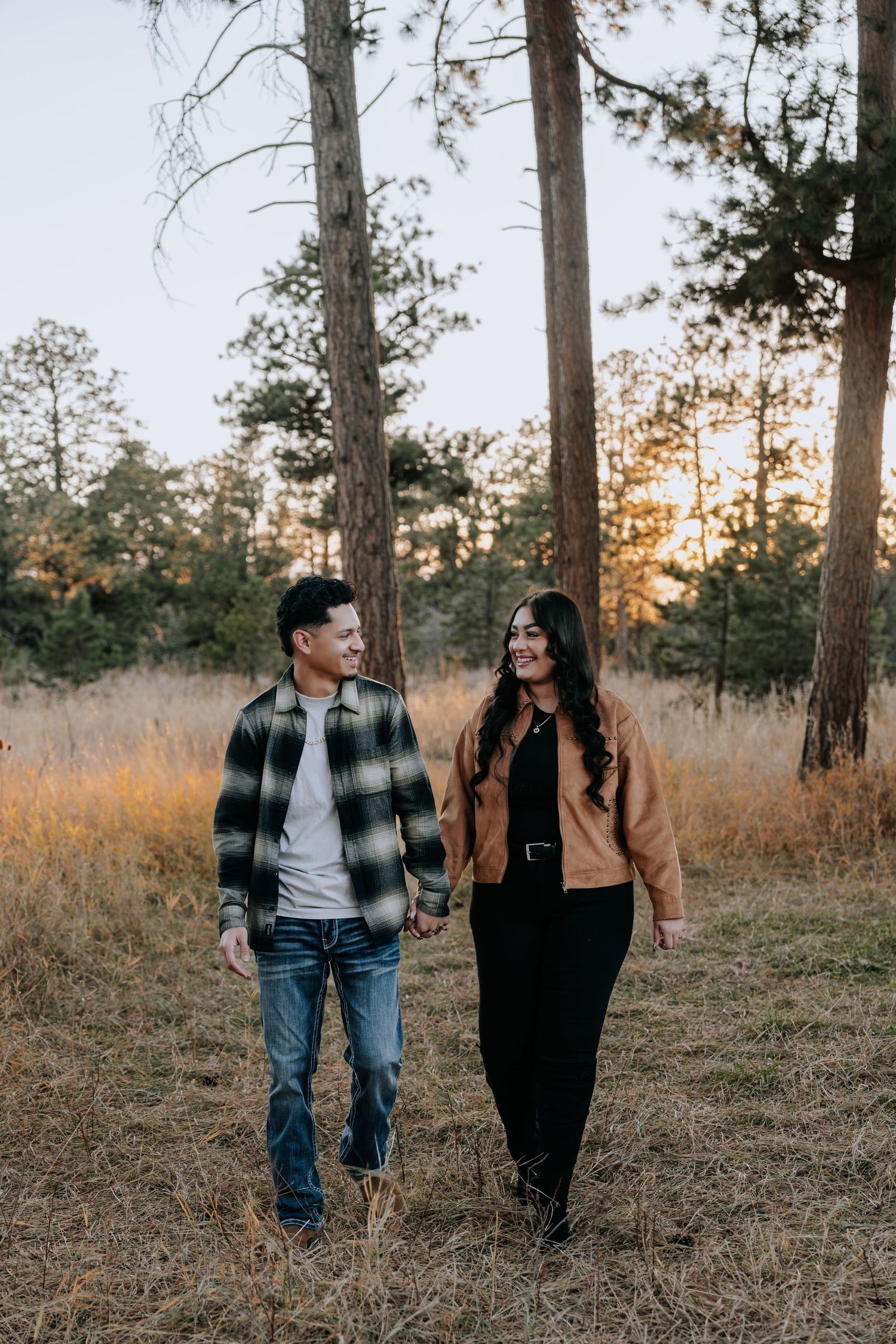 Happy couple holding hands walking through a golden grass field with pine trees at sunset.