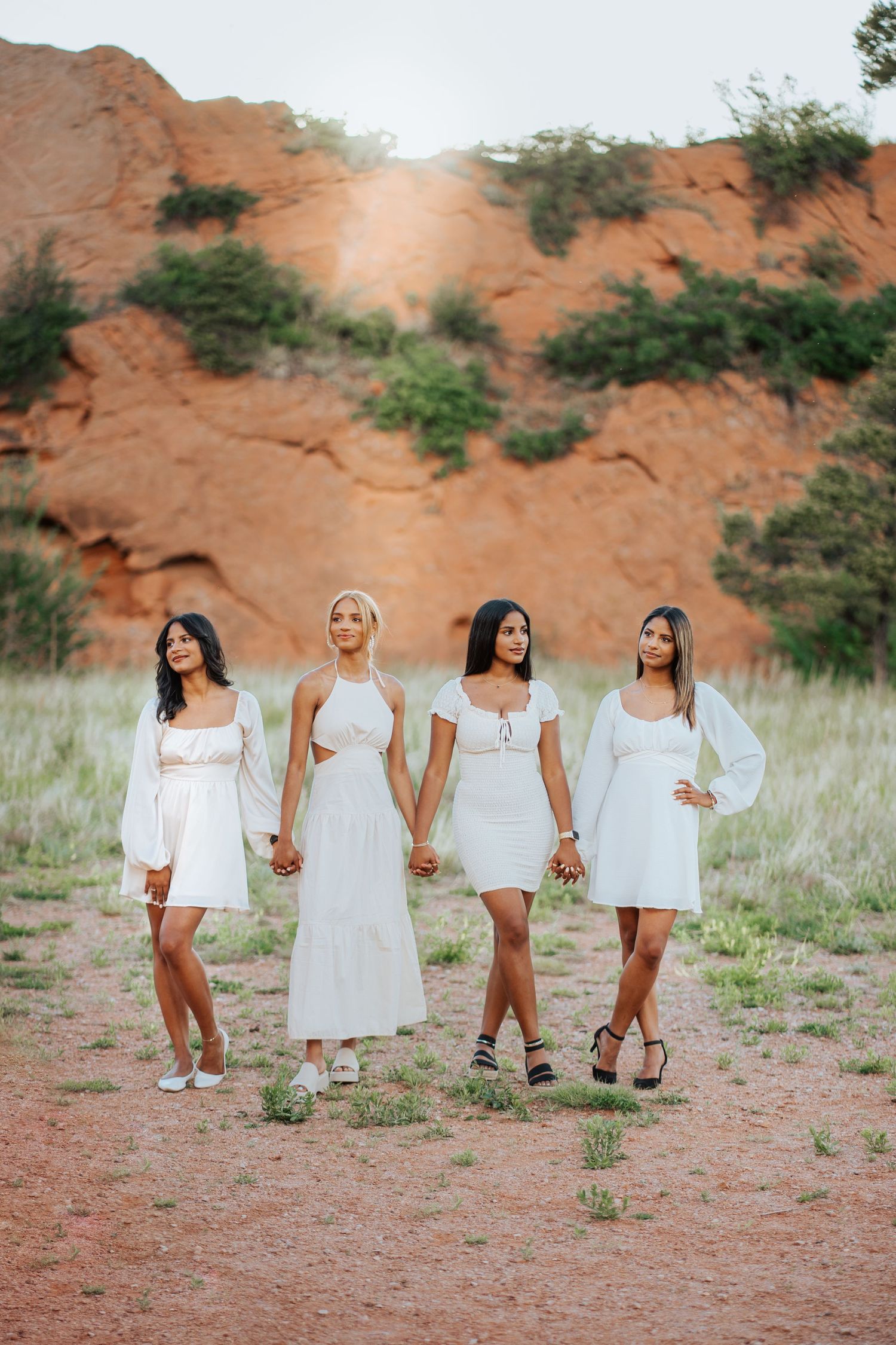 Four women in white dresses hold hands walking through a red rock landscape in Colorado.
