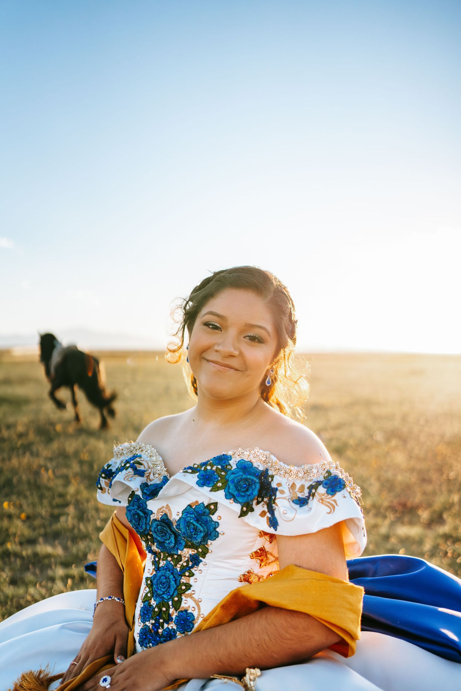 Smiling quinceañera in white blue floral off-shoulder gown seated in sunny field with horse.