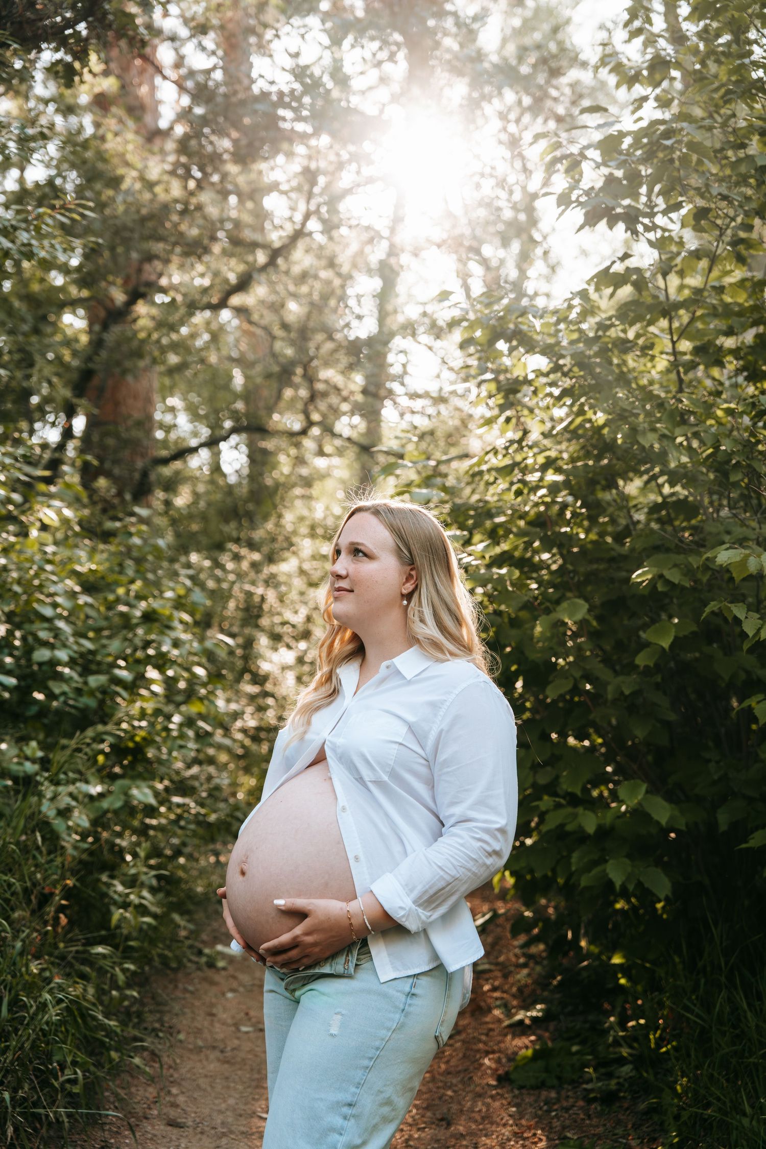 Pregnant woman in white shirt and jeans holds belly on a sunny nature path.