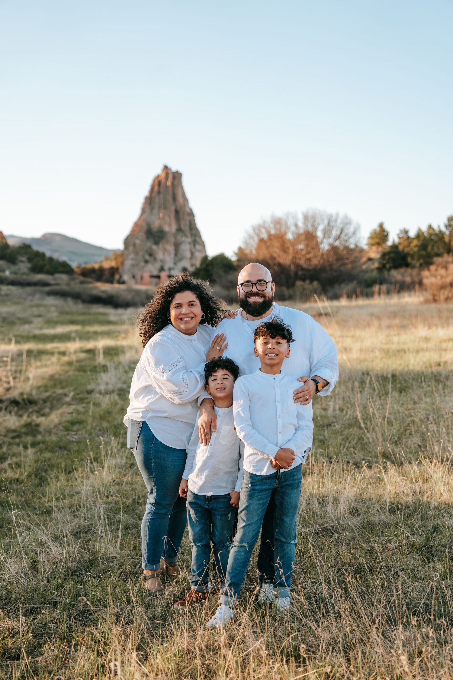 Happy family of four in matching white tops and jeans posing in field with rock formation backdrop.