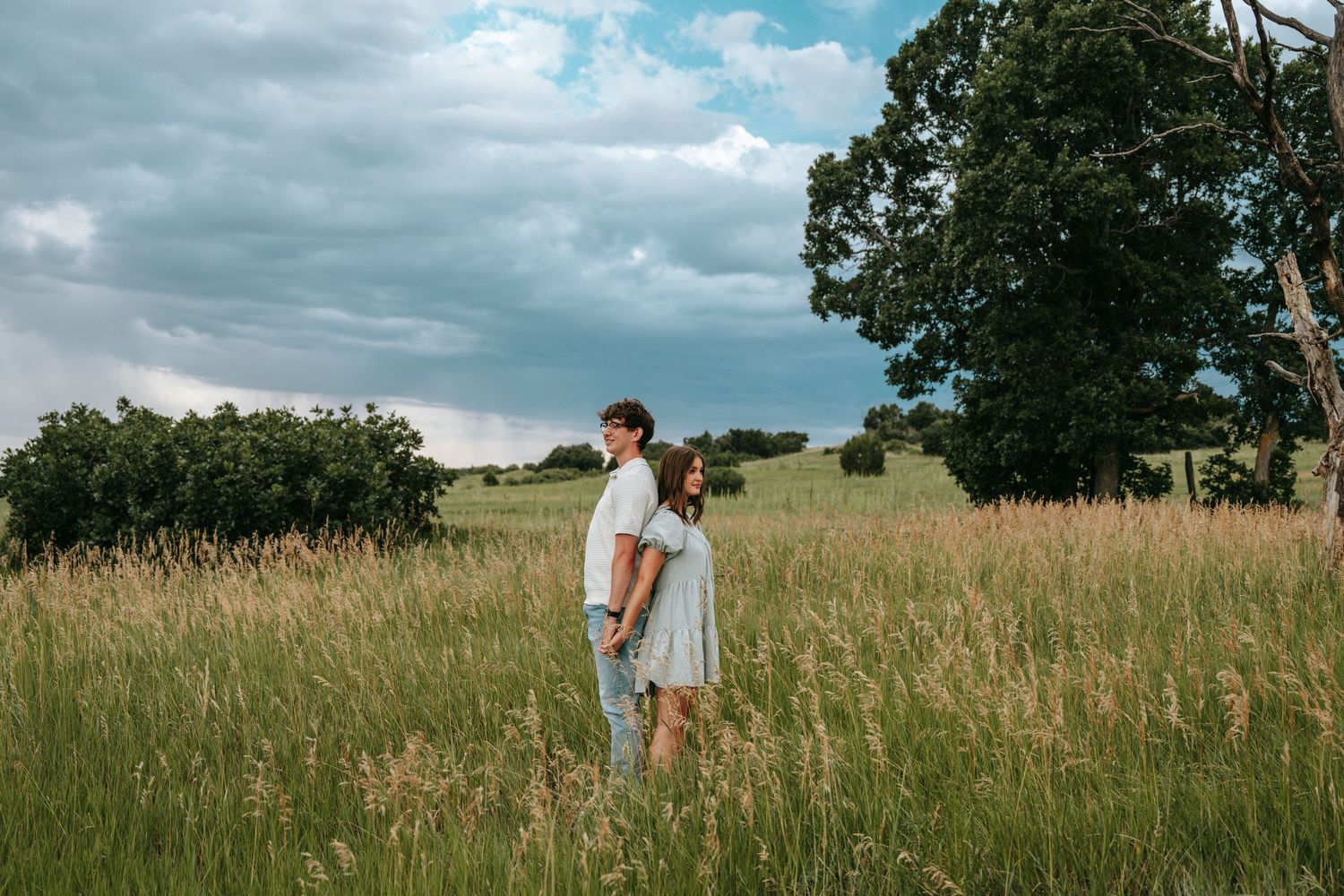 Couple standing back to back in a tall grass meadow with a dramatic cloudy sky backdrop.