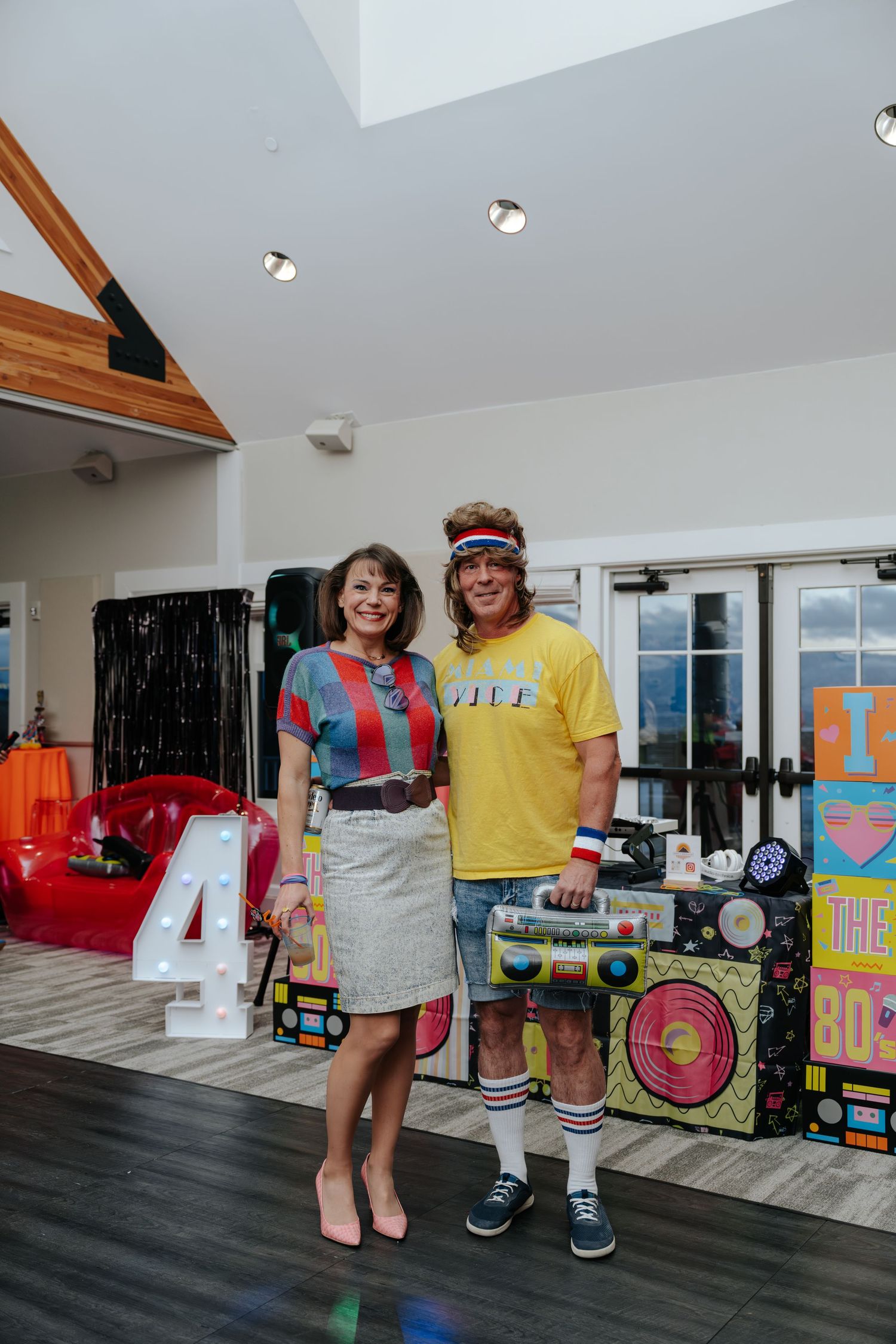 Two people in 80s themed costumes pose at a retro decade birthday party with boombox and colorful decorations.