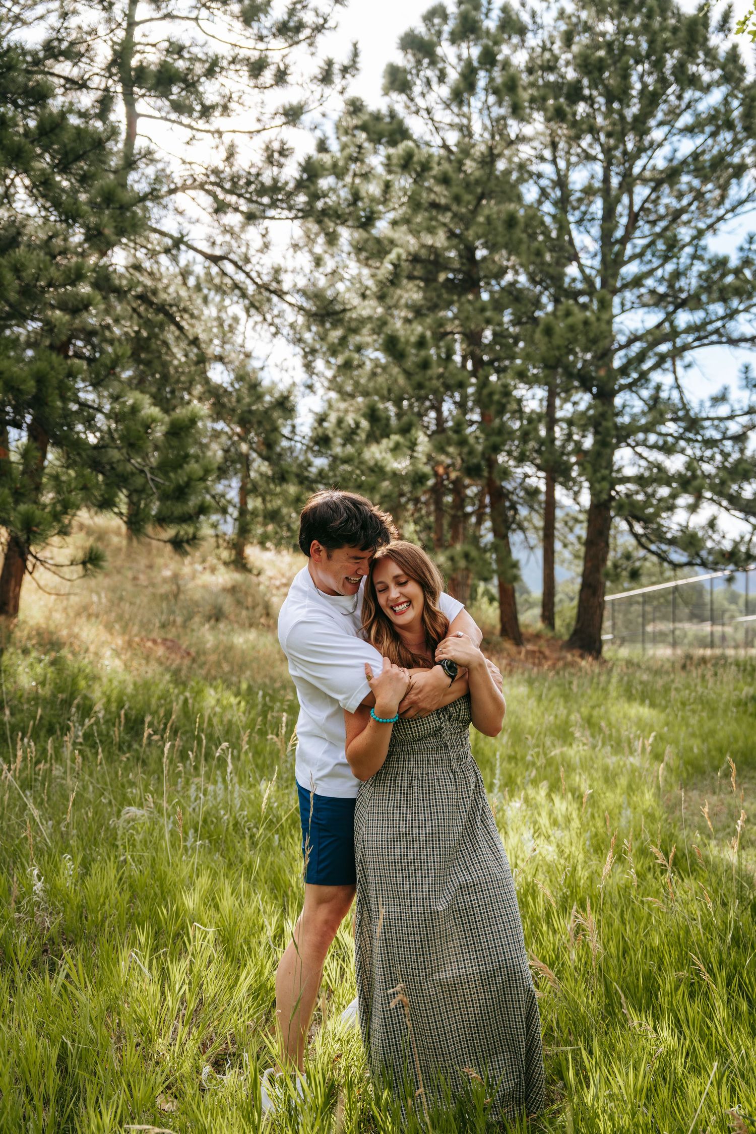 Couple embracing and laughing in a lush green meadow surrounded by tall pine trees during an outdoor engagement session.