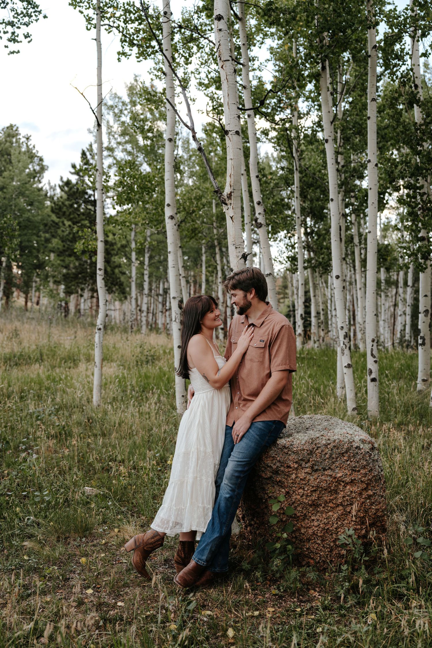 Couple sharing a romantic moment by a boulder in an aspen forest, woman in white dress and cowboy boots.