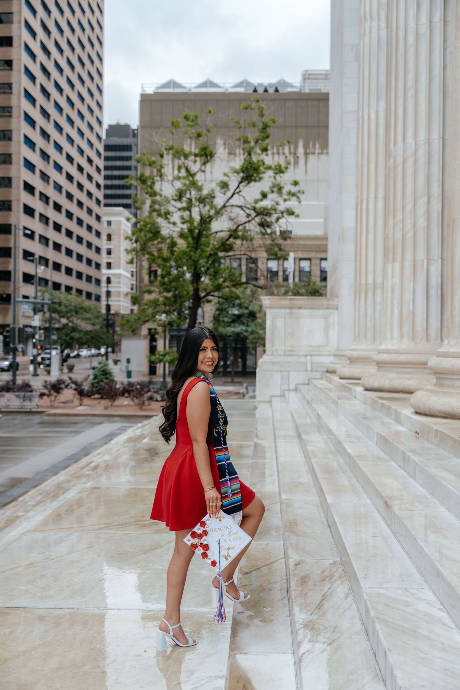 Young woman in red dress holding decorated graduation cap poses on marble steps with city buildings behind her.