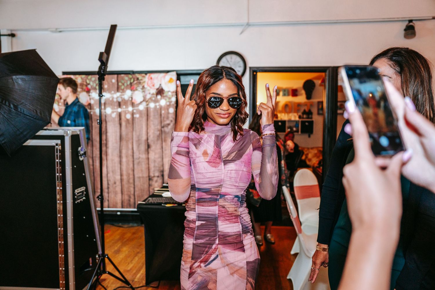 Woman in purple patterned dress posing with sunglasses while guests photograph her at party.