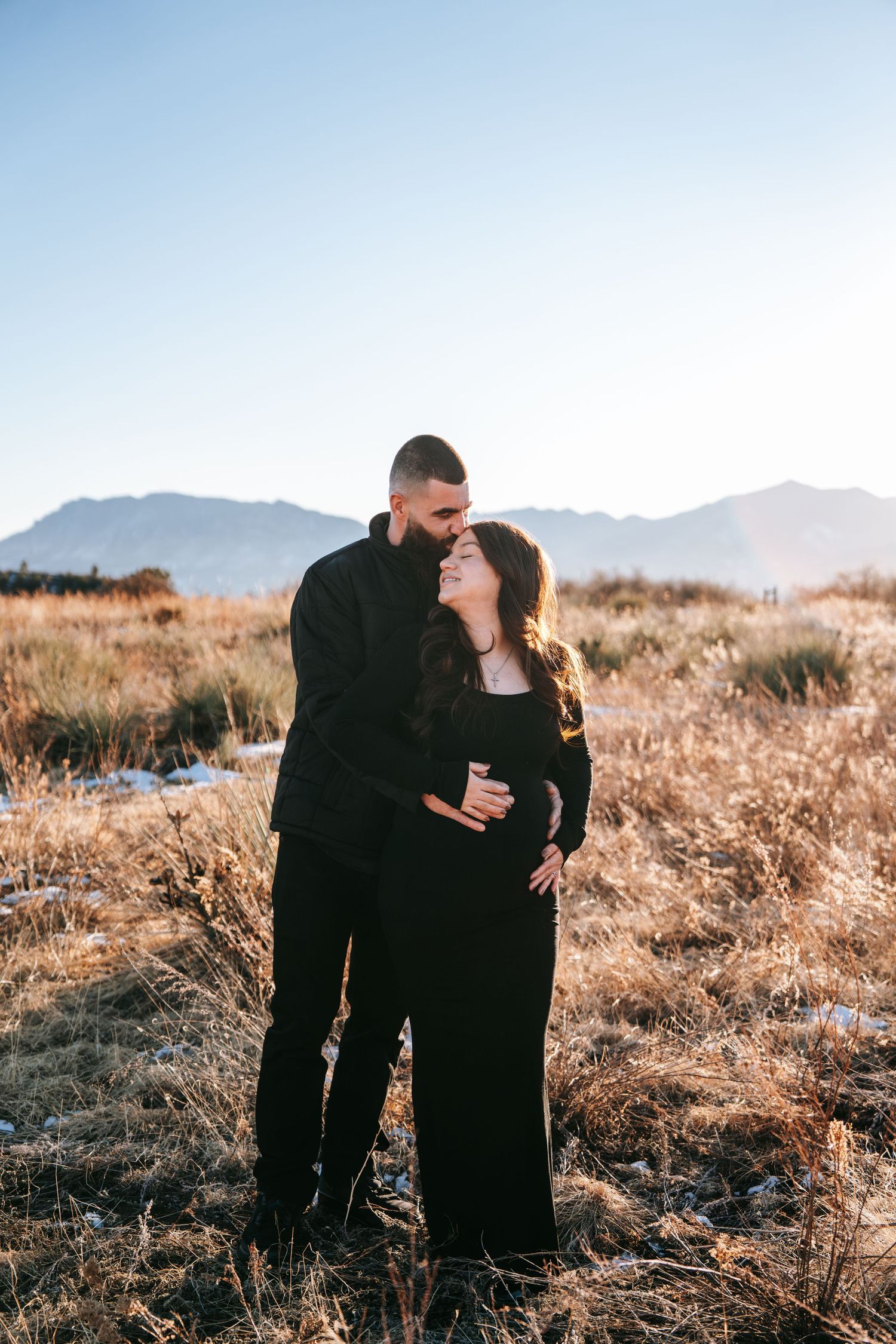 Couple in matching black outfits sharing tender moment during outdoor maternity photoshoot with mountain backdrop.
