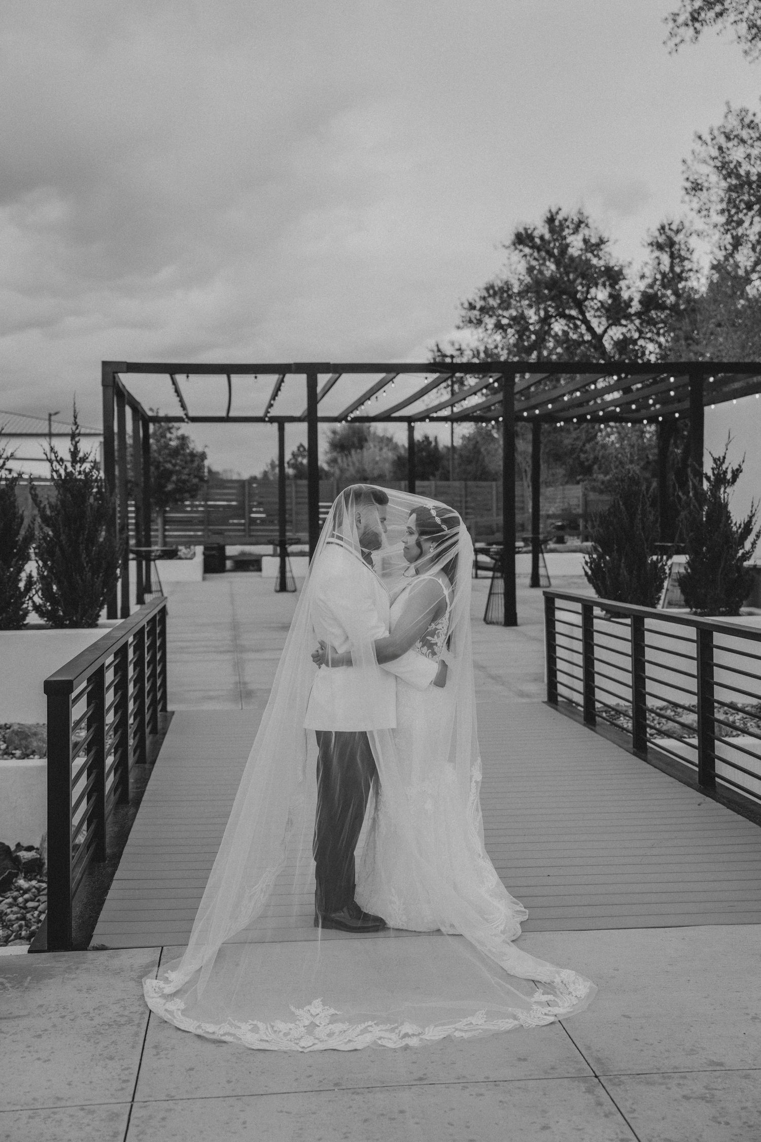 Black and white photo of bride and groom sharing a kiss under cathedral veil on outdoor venue bridge.