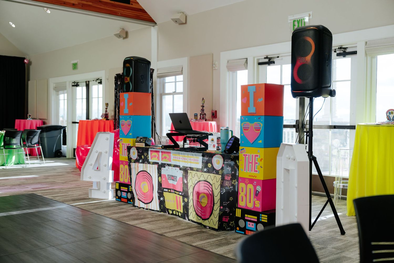 Colorful 80s-themed DJ booth setup in a bright event hall with neon decorations and speaker stands.