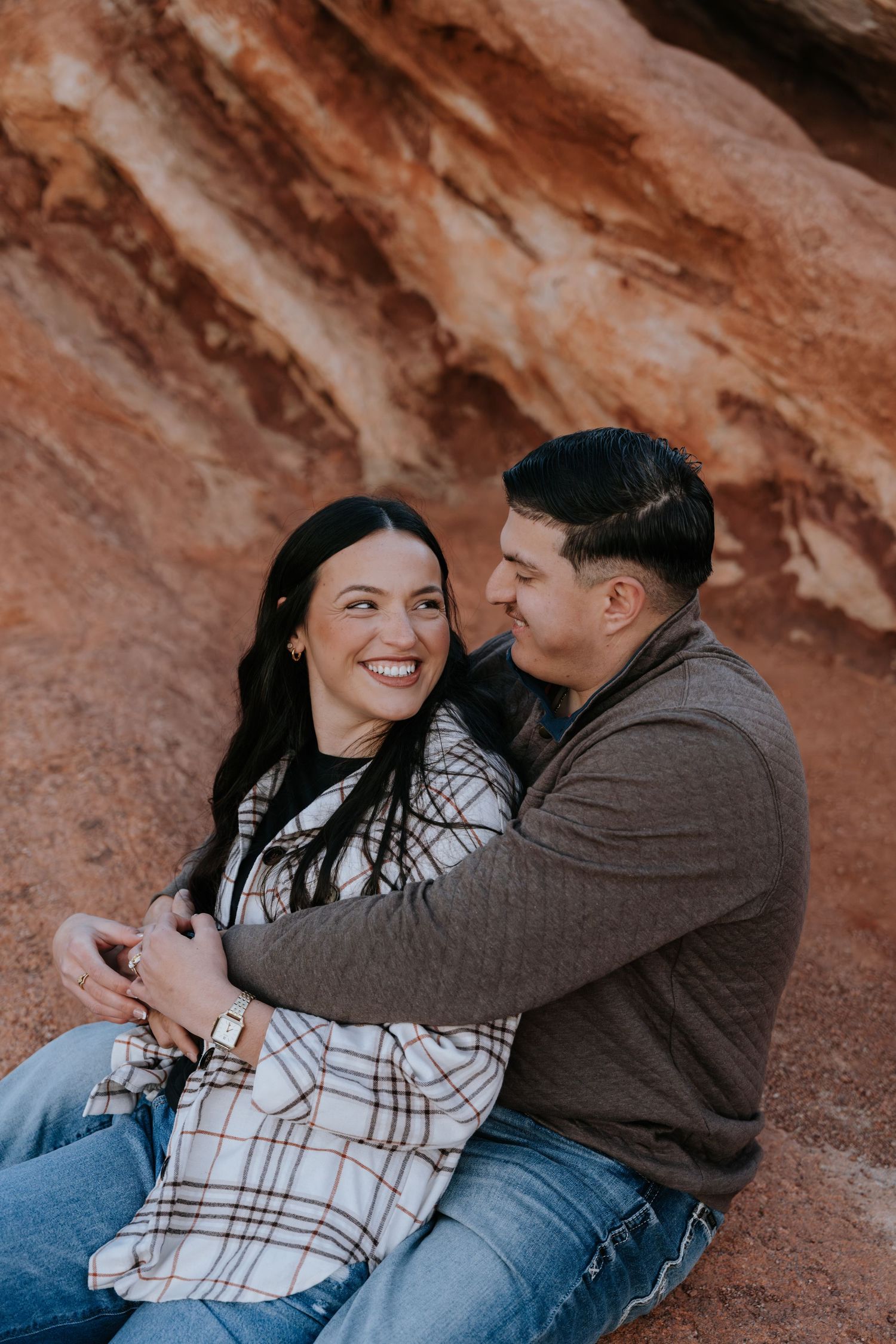 Couple sitting and embracing against vibrant red rock wall during romantic outdoor engagement session.