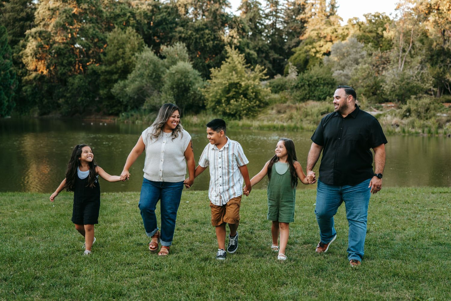 Happy family of five holding hands and walking together in a park by a lake.