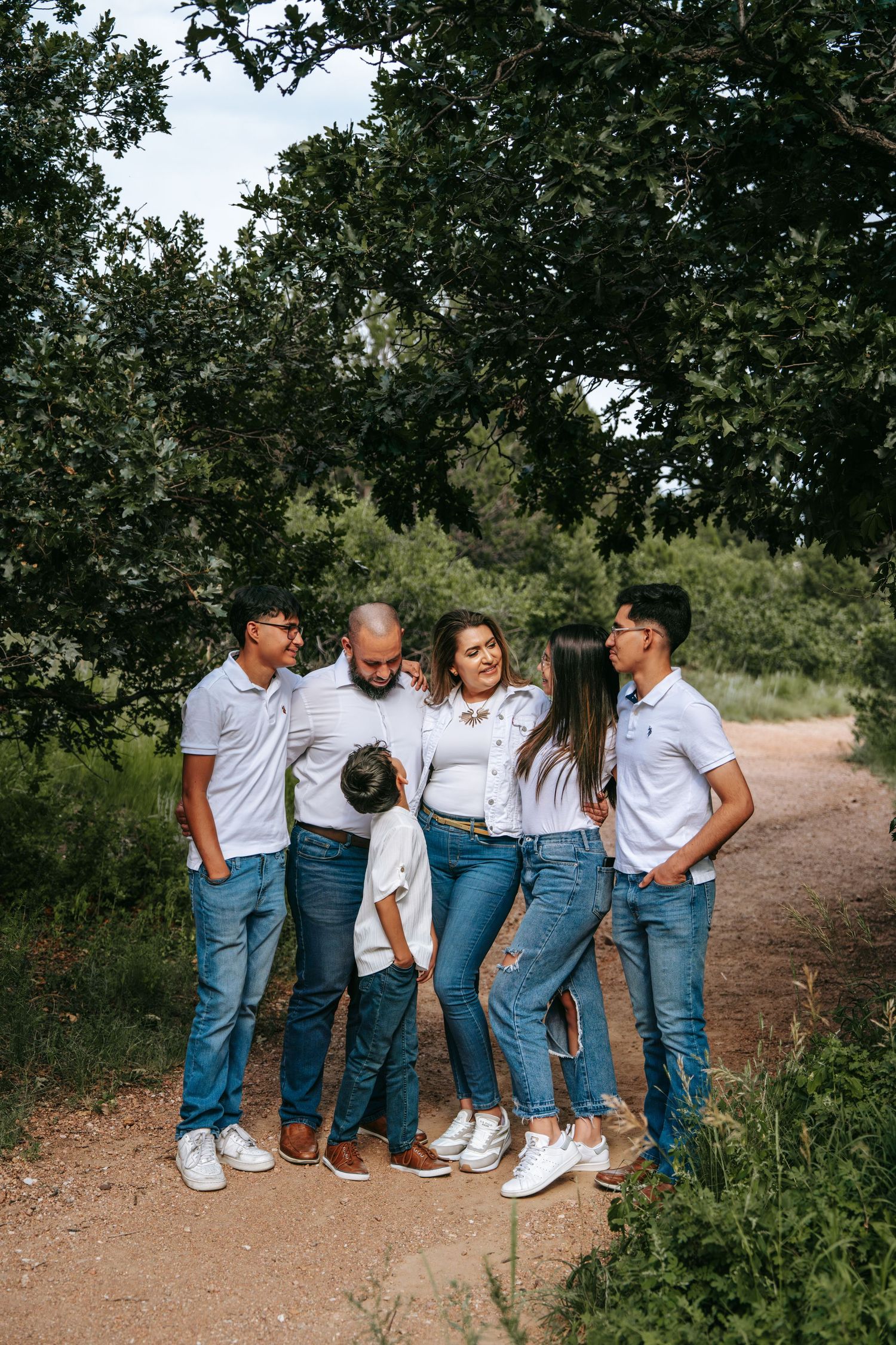 Family of five in matching white shirts and jeans posing together on a dirt path surrounded by lush green trees.