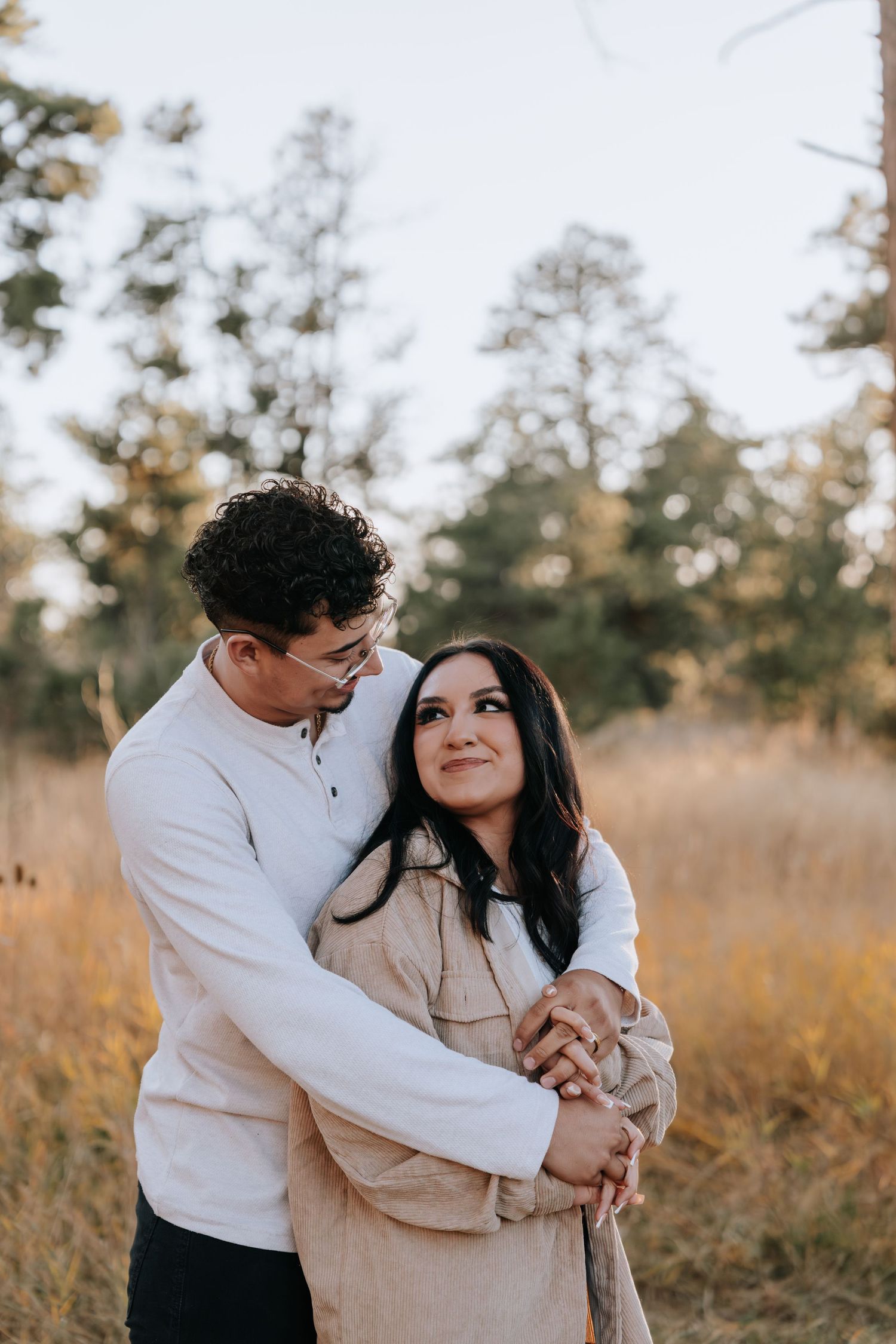 Couple embracing in a golden meadow surrounded by tall pine trees in soft evening light.