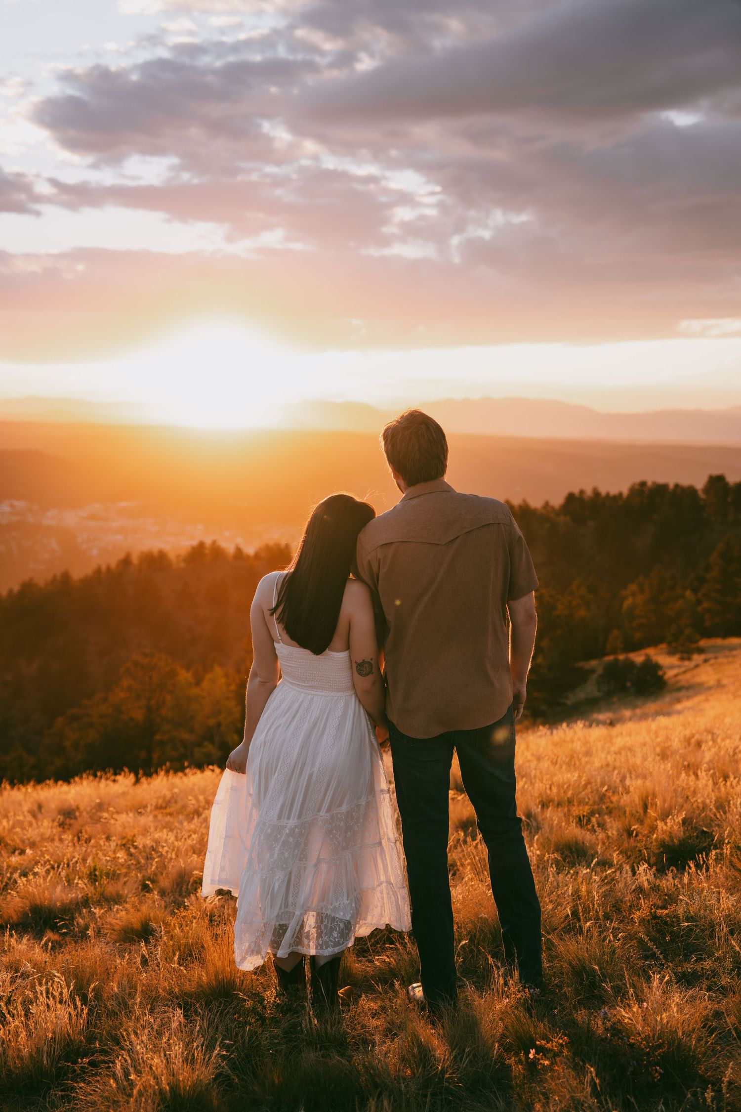 Couple embracing while watching a golden sunset over mountain landscape, woman in white dress, romantic outdoor photography.