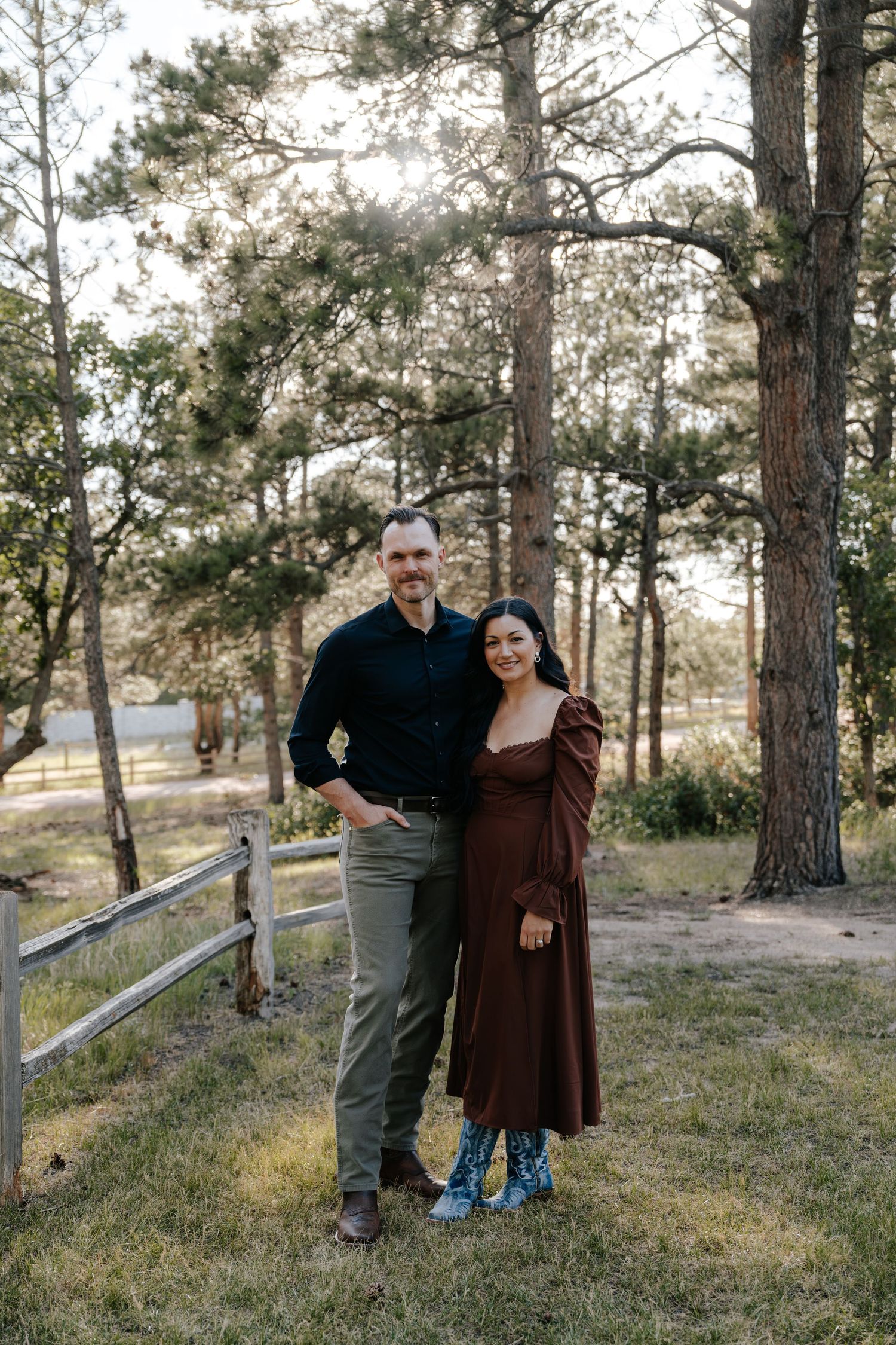 Couple smiling together outdoors near a wooden fence surrounded by tall pine trees in a forest setting.