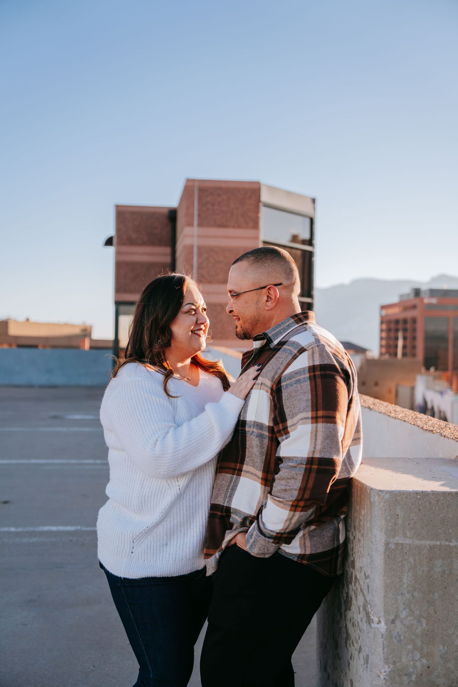 Smiling couple facing each other on a rooftop at golden hour with urban buildings in background.