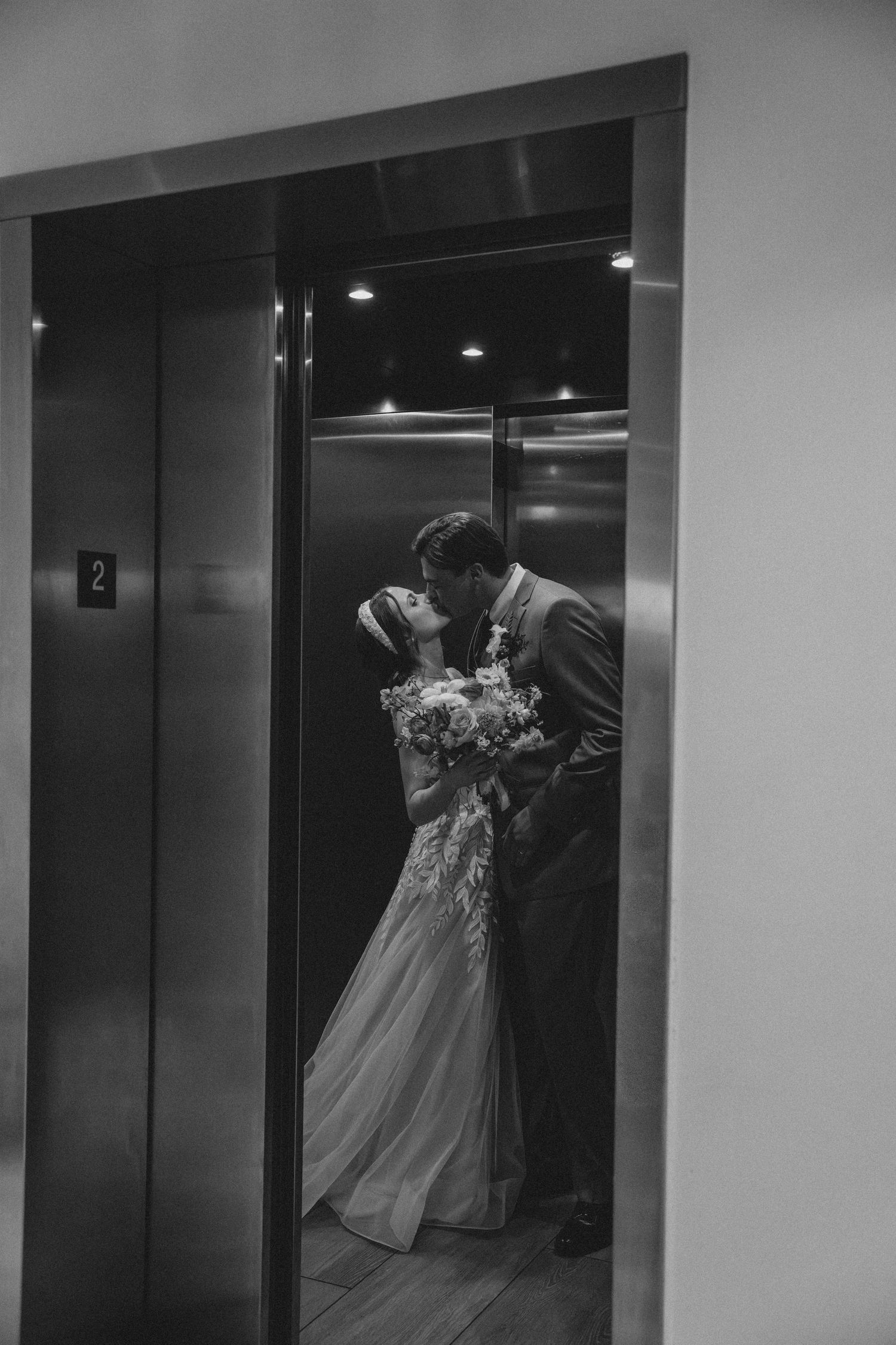Black and white photo of bride and groom sharing a kiss inside an elevator.