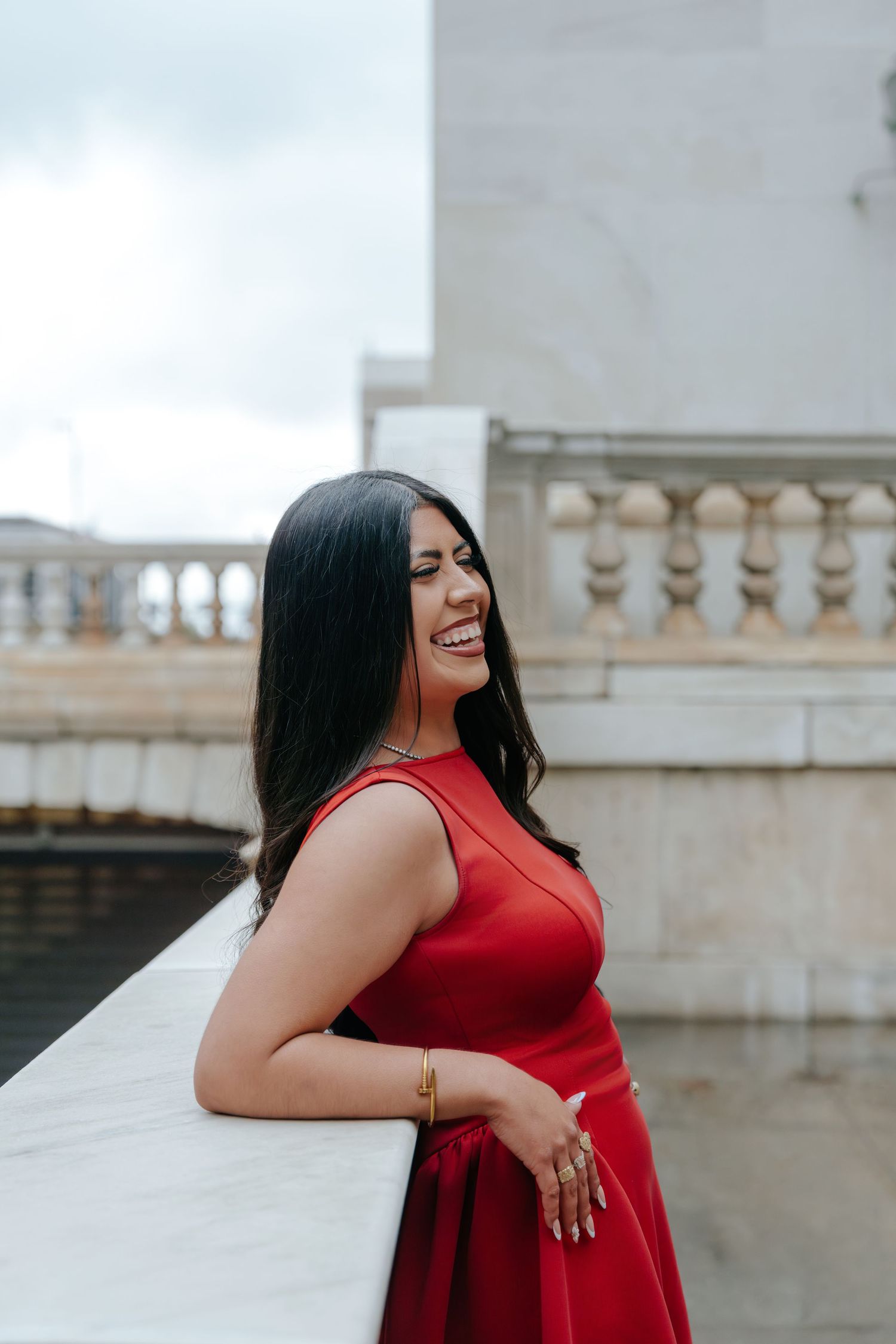 Laughing woman in red dress leaning on white marble railing near ornate stone balustrade.