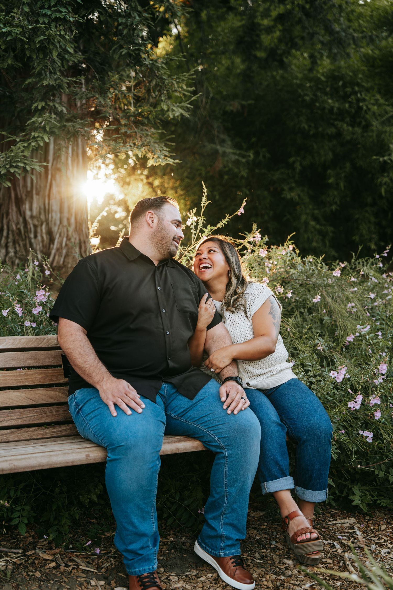A laughing couple sits together on a wooden bench surrounded by blooming flowers at sunset.
