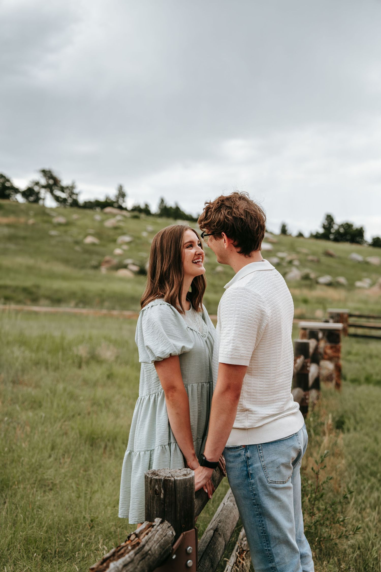 Romantic couple holding hands and gazing at each other by a rustic wooden fence in a green pasture.