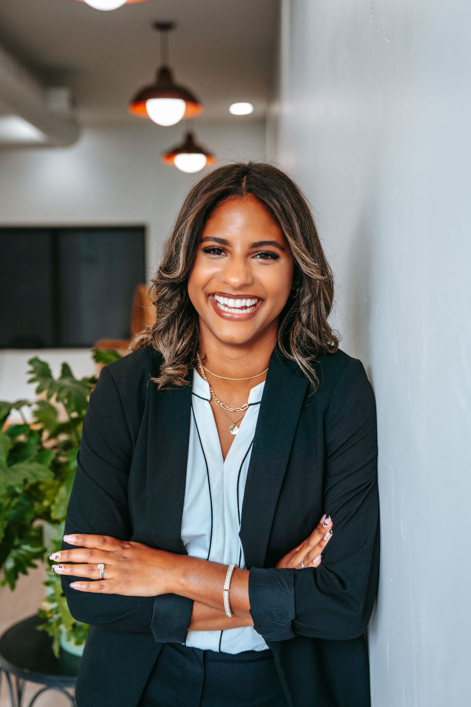 Confident young businesswoman in black blazer smiling with arms crossed in a modern office environment.