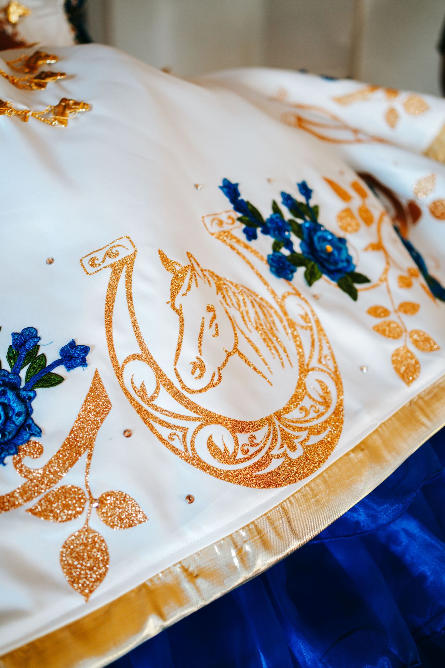 White quinceañera dress with gold horseshoe and horse embroidery with blue floral accents.