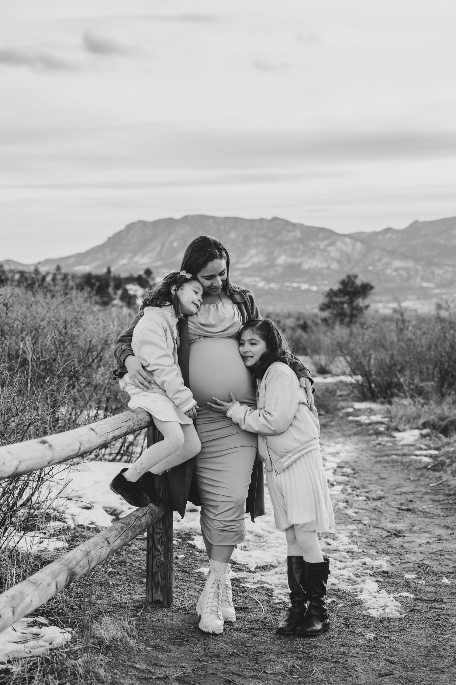 Black and white photo of pregnant mother with two daughters hugging her baby bump near a wooden fence outdoors.