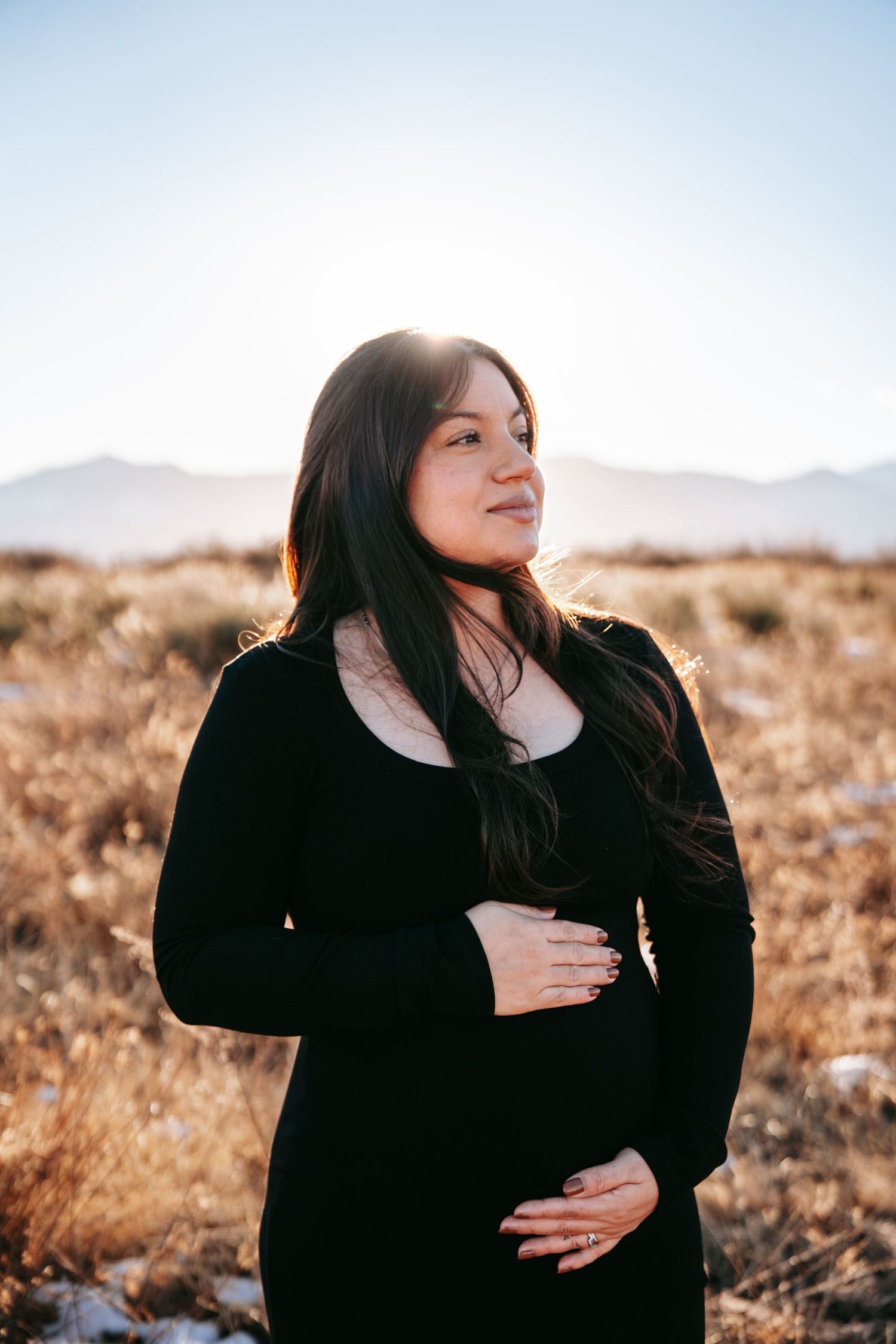 Glowing pregnant woman in black dress holding baby bump outdoors in desert field with soft sunlight.