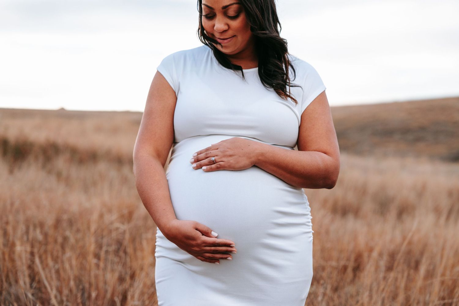 Pregnant woman in white dress looks down tenderly at her baby bump in a golden grass field.