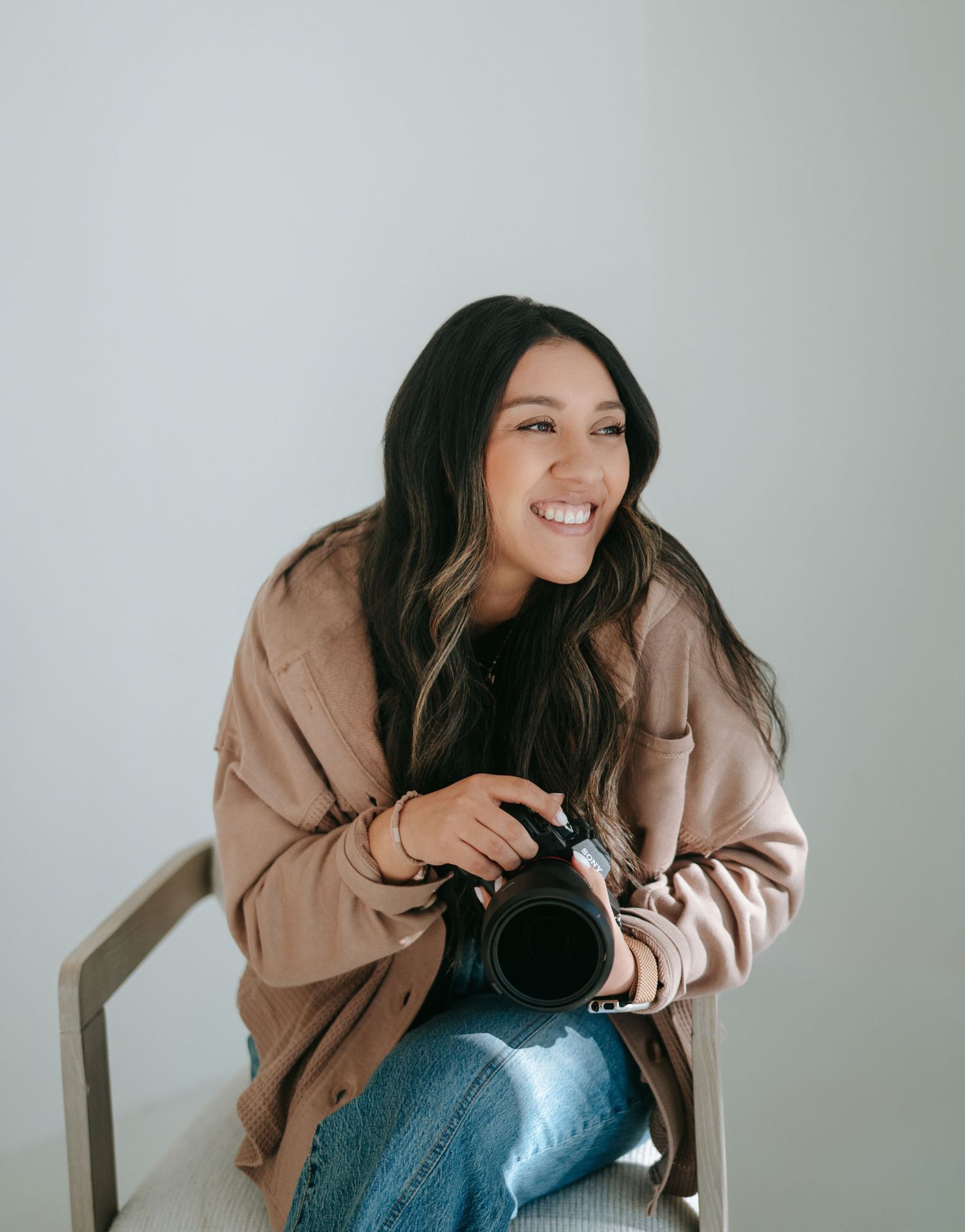 Smiling woman with long dark hair holds a camera while seated, wearing a beige coat.