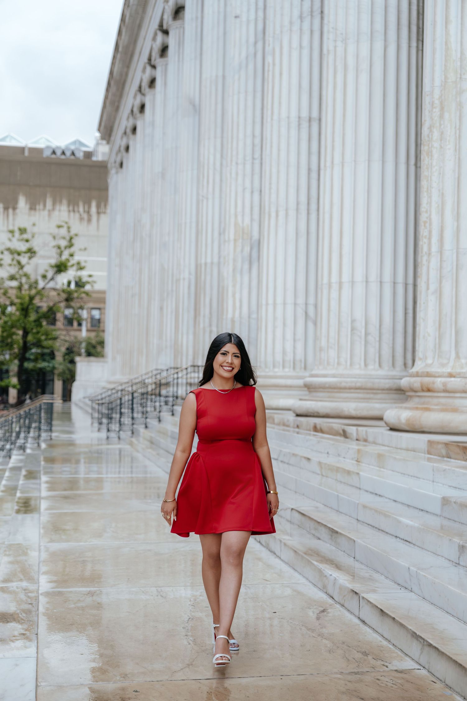 Smiling young woman in red sleeveless dress walks along wet marble plaza beside towering classical columns.