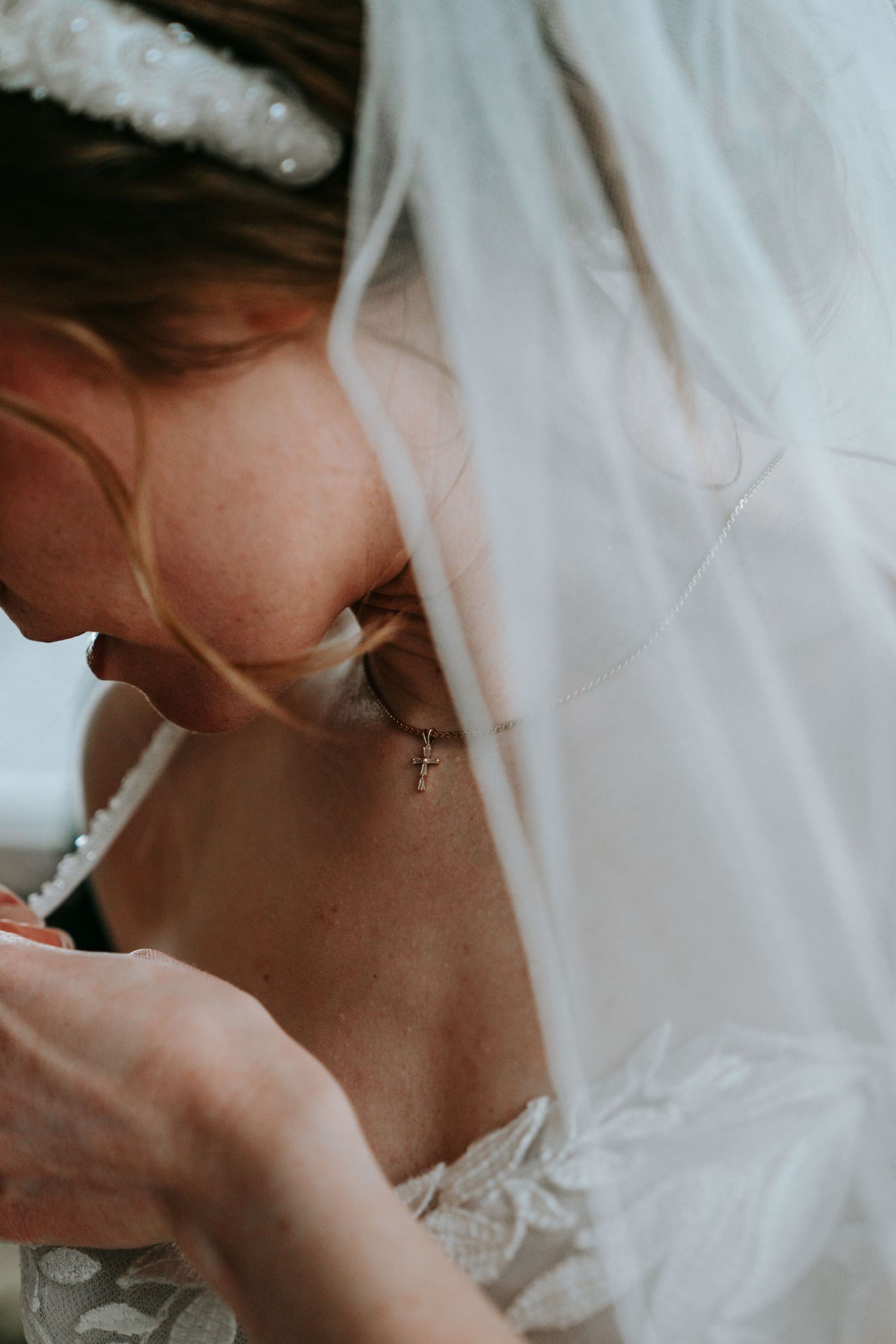 Close-up of bride wearing a delicate cross necklace and pearl headband with veil draped over her face.
