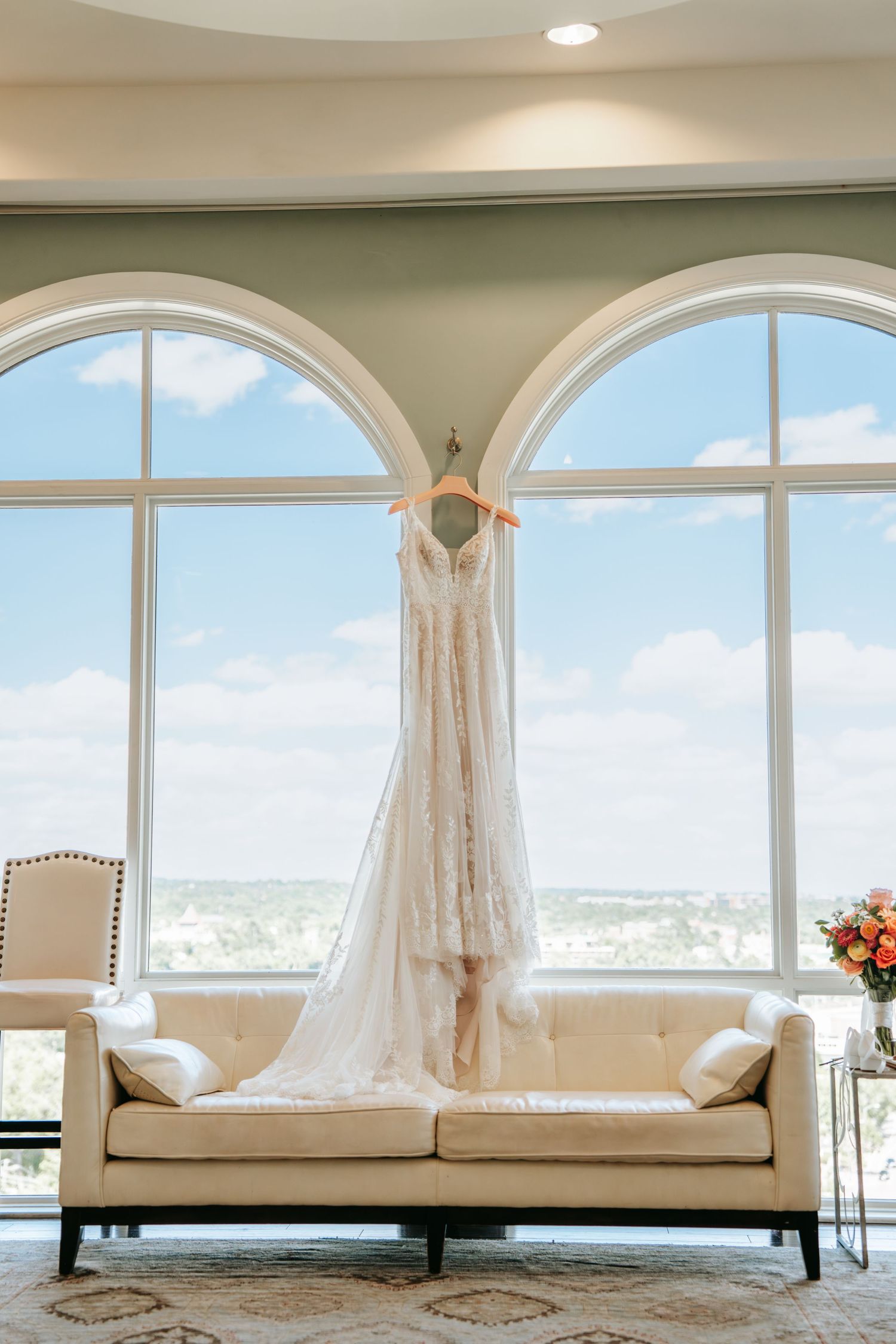 Elegant lace bridal gown displayed on wooden hanger above cream sofa in front of large arched windows.