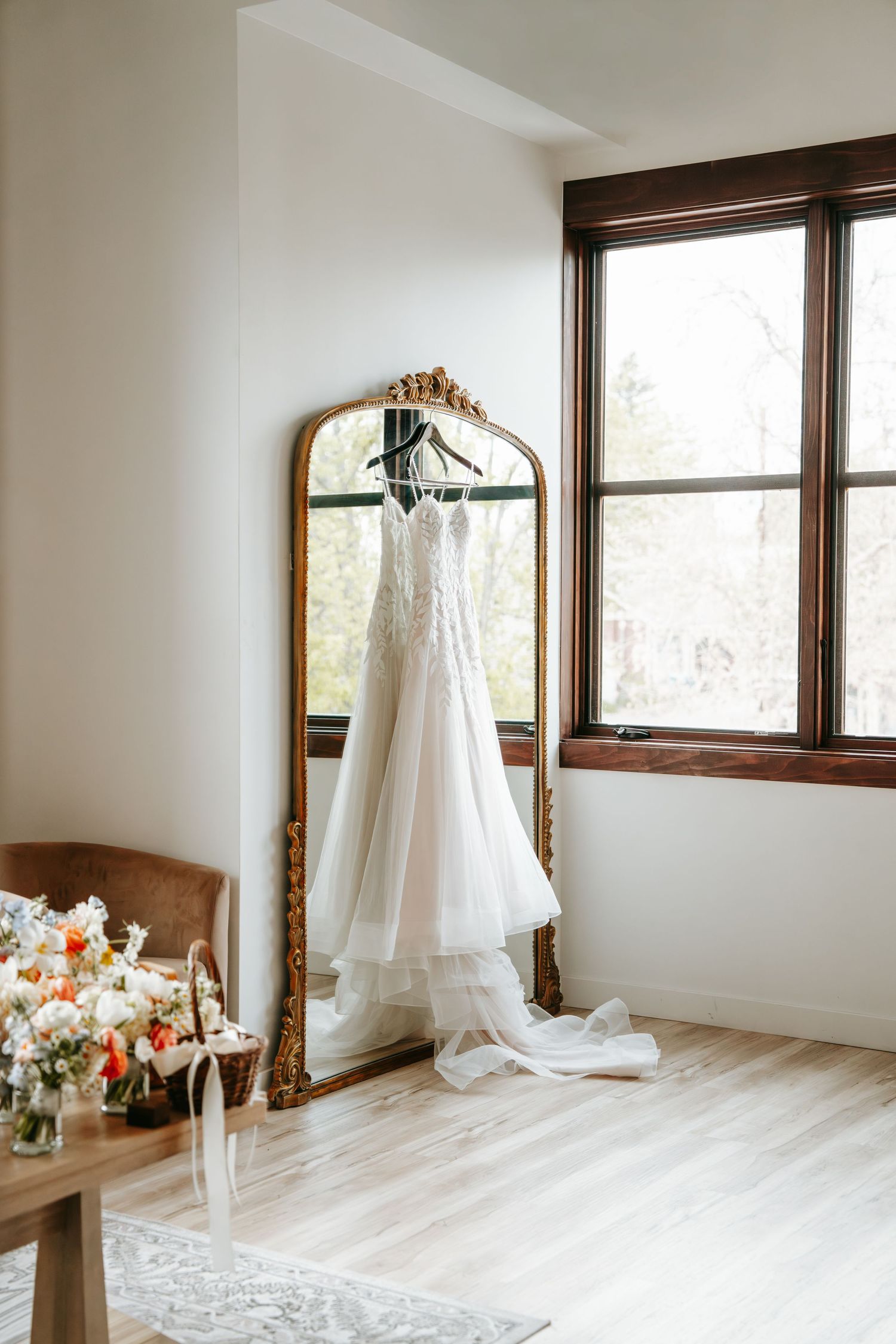 White wedding gown hanging on ornate gold mirror in bright bridal suite with hardwood floors.