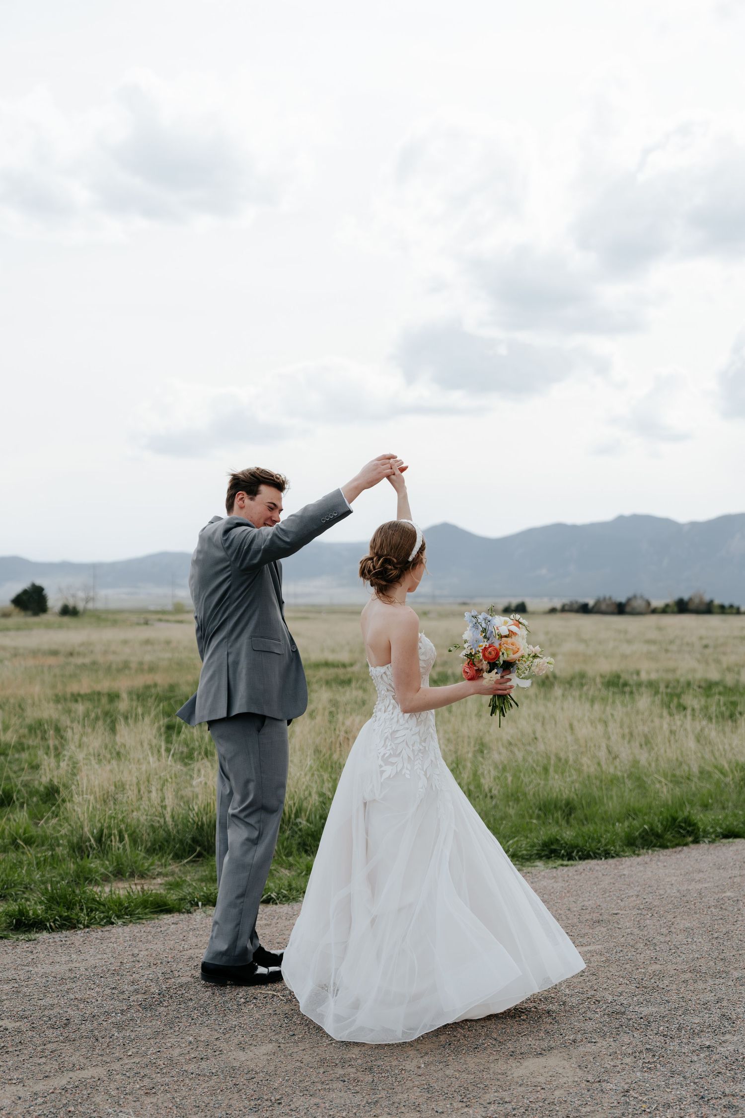 Groom in gray suit twirls bride in white lace wedding dress on a scenic mountain path.