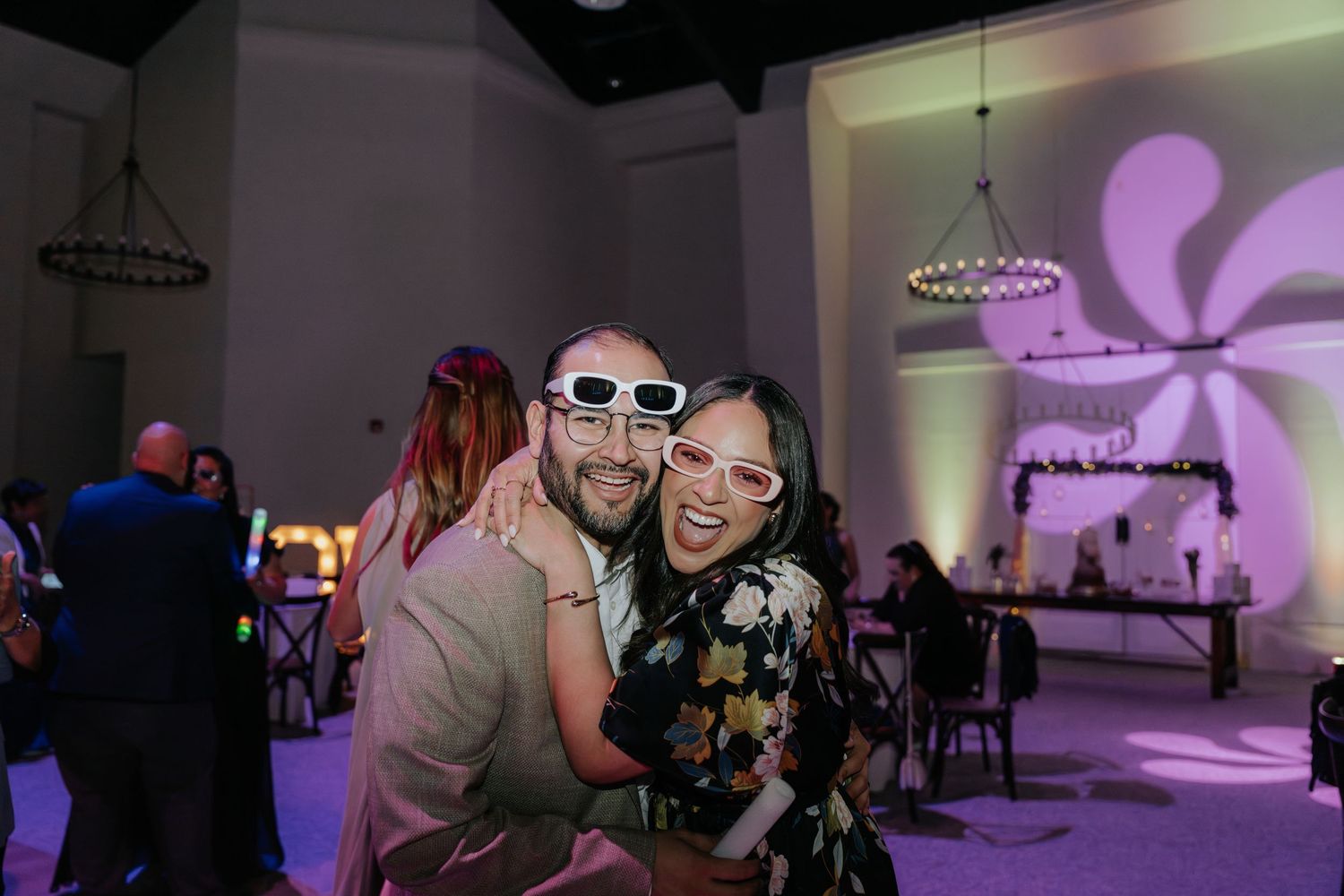 Two people wearing white sunglasses smile and hug at an elegant wedding reception with purple lighting.