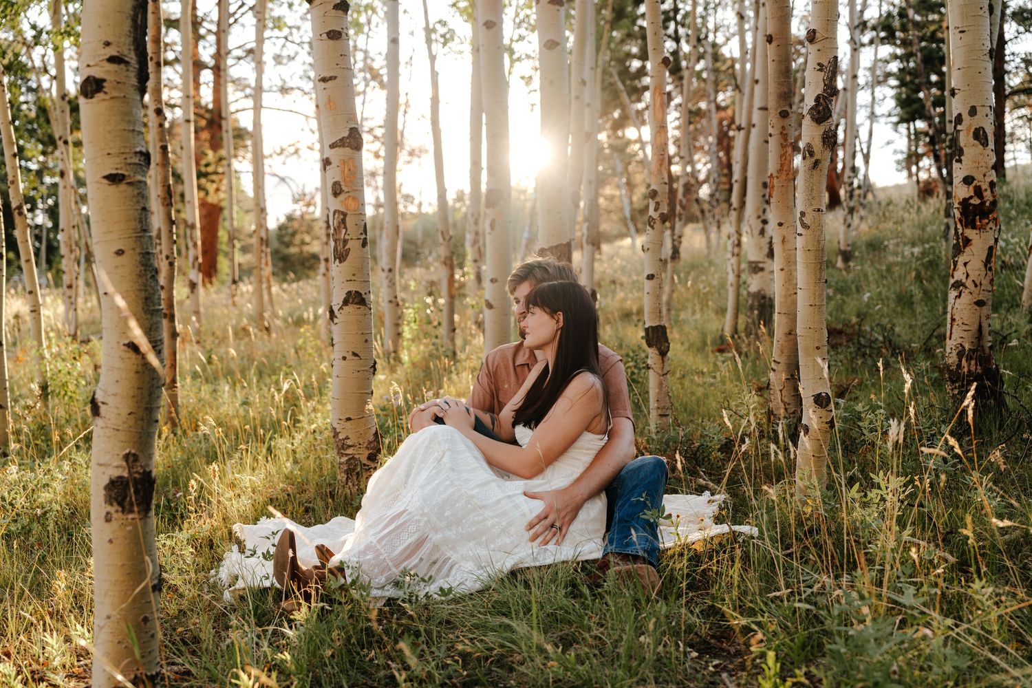 Couple sitting together in a sunlit aspen forest, surrounded by tall white birch trees and green grass.