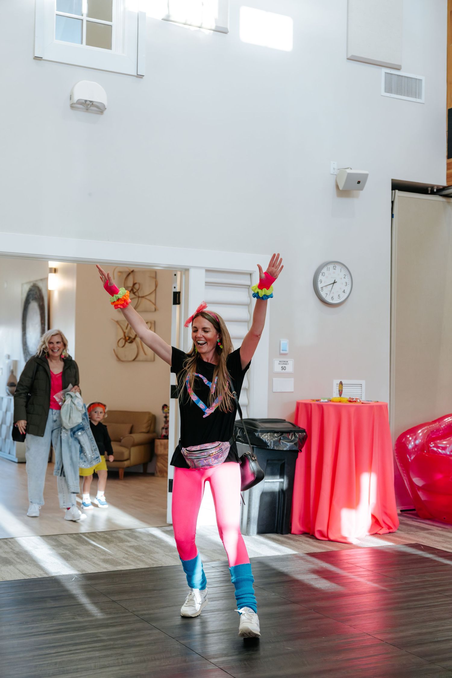 Woman in colorful 80s outfit with neon pink leggings, fanny pack, and leg warmers dancing at a retro-themed event.