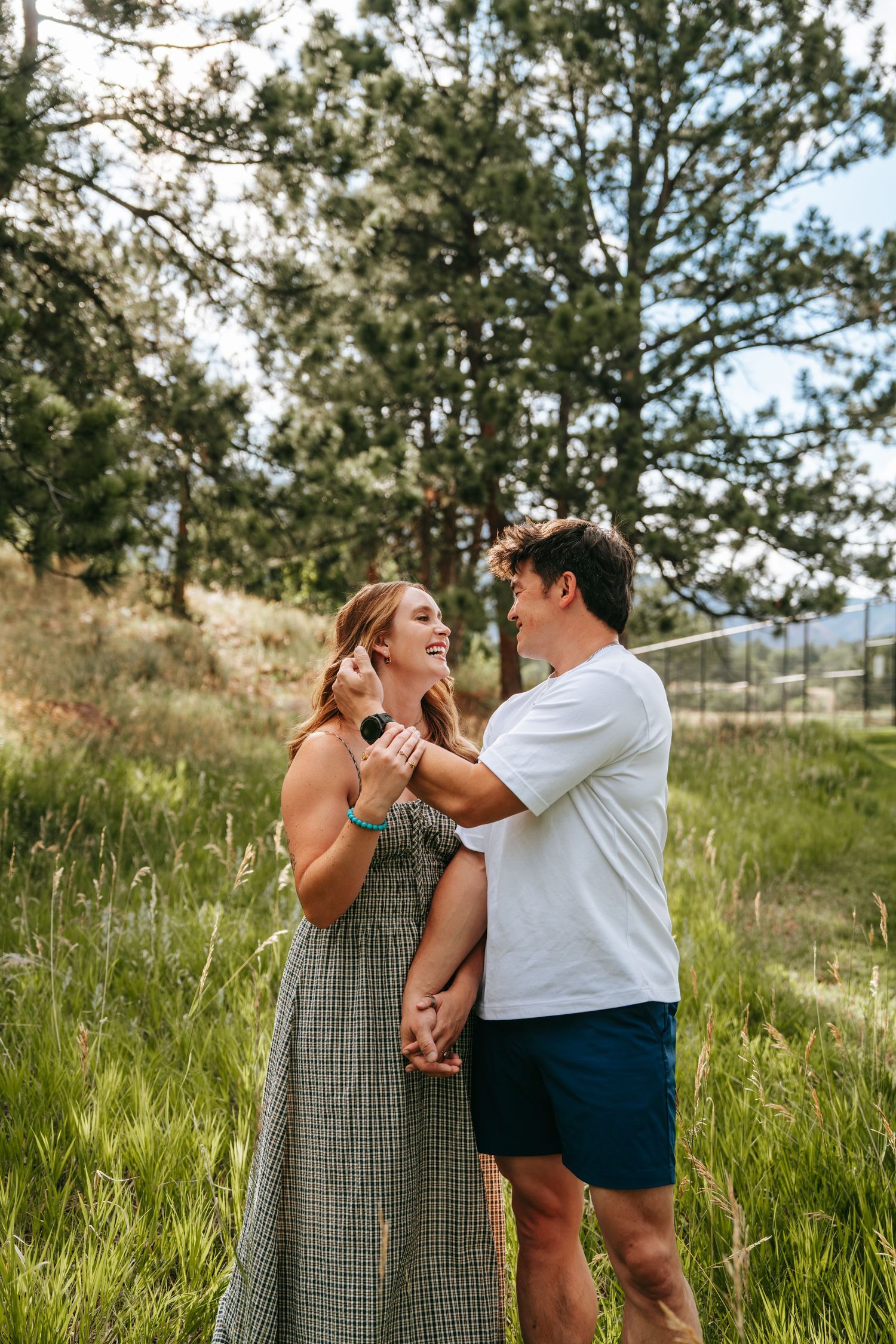 Happy couple embracing and smiling in a lush green meadow surrounded by tall pine trees on a sunny day.