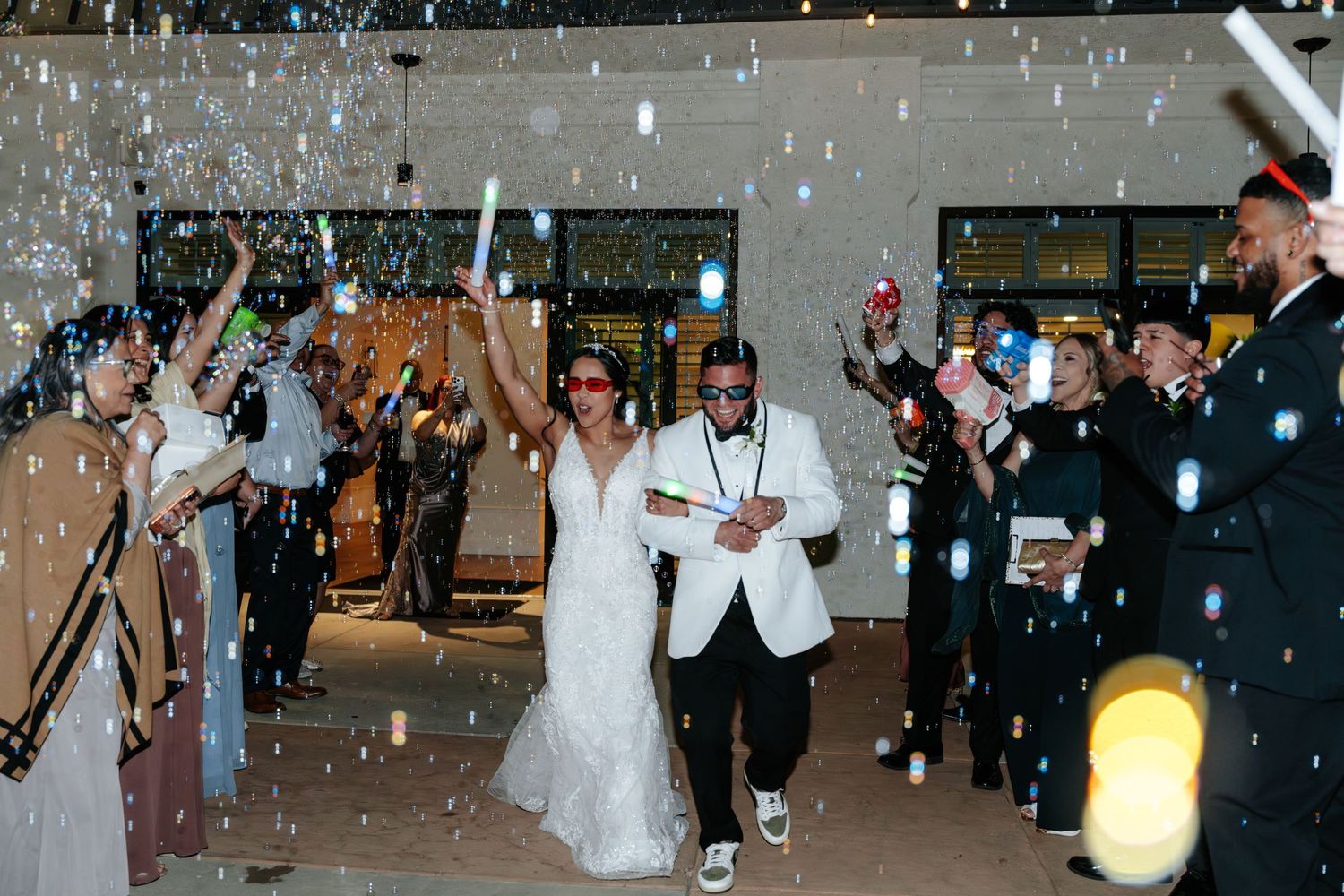 Bride and groom exit wedding reception through bubble send-off, guests cheering with glow sticks at night.