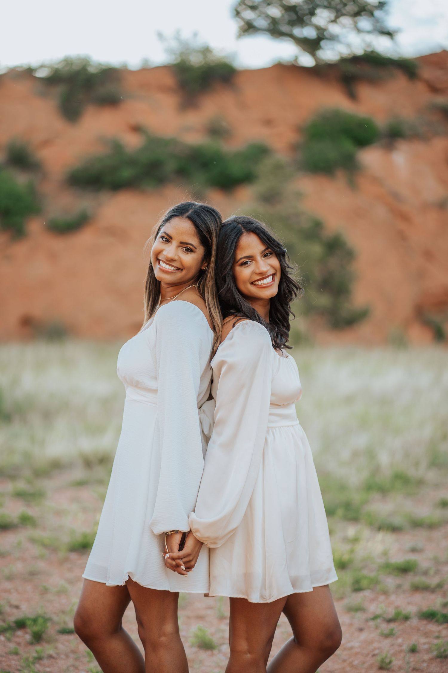 Two young women in white dresses smiling back-to-back against red rock formation backdrop.
