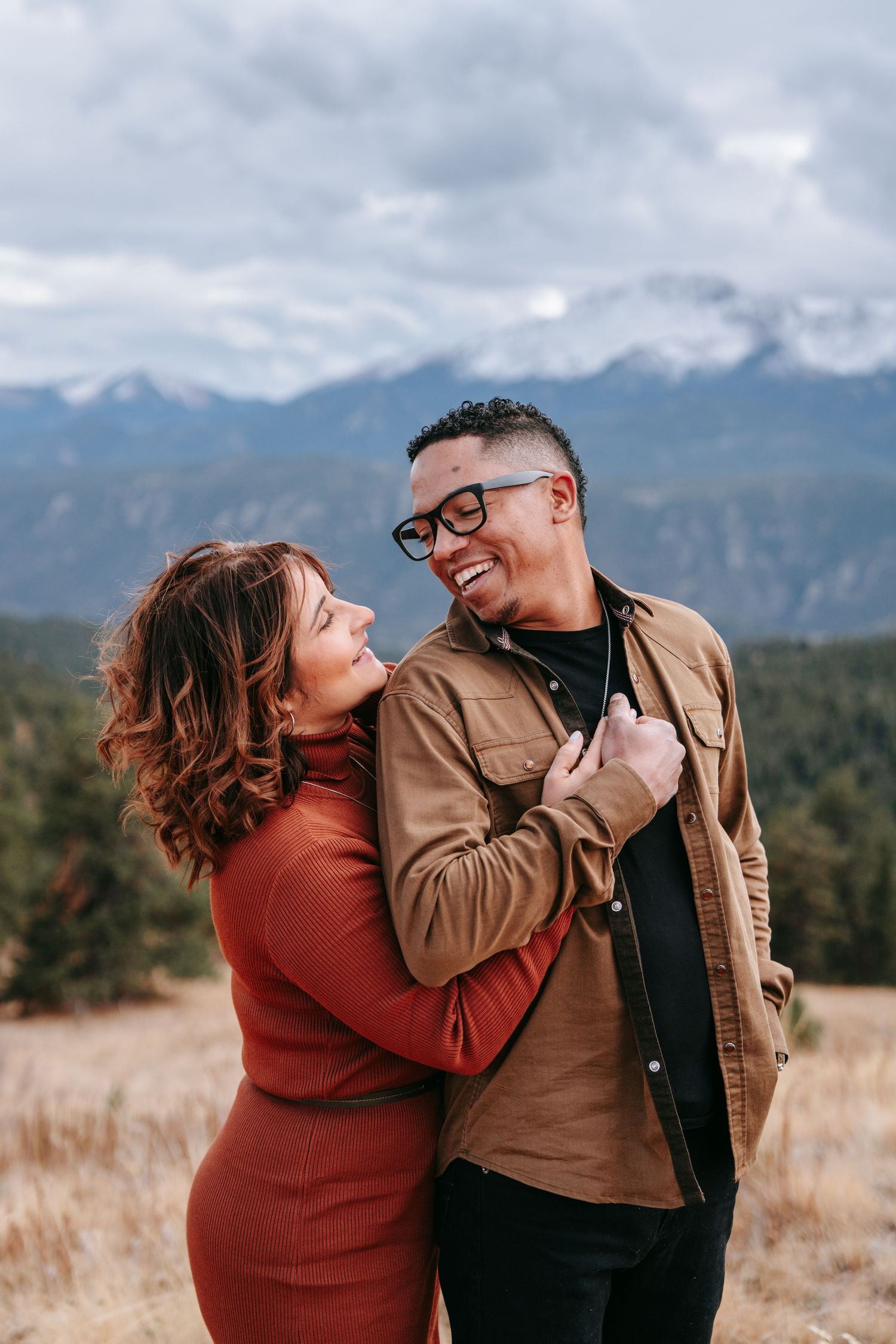 Happy couple laughing together in a mountain meadow, woman in rust dress holding mans jacket lapels.