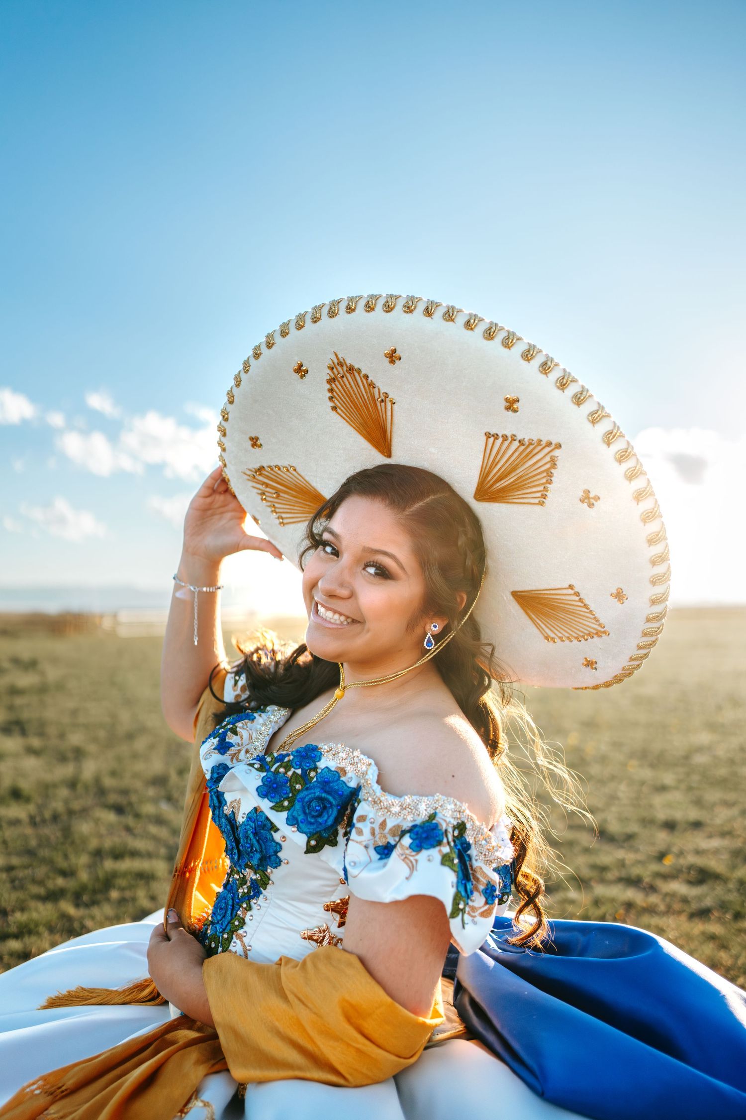 Young woman in quinceañera dress smiling while holding white gold sombrero under blue sky.
