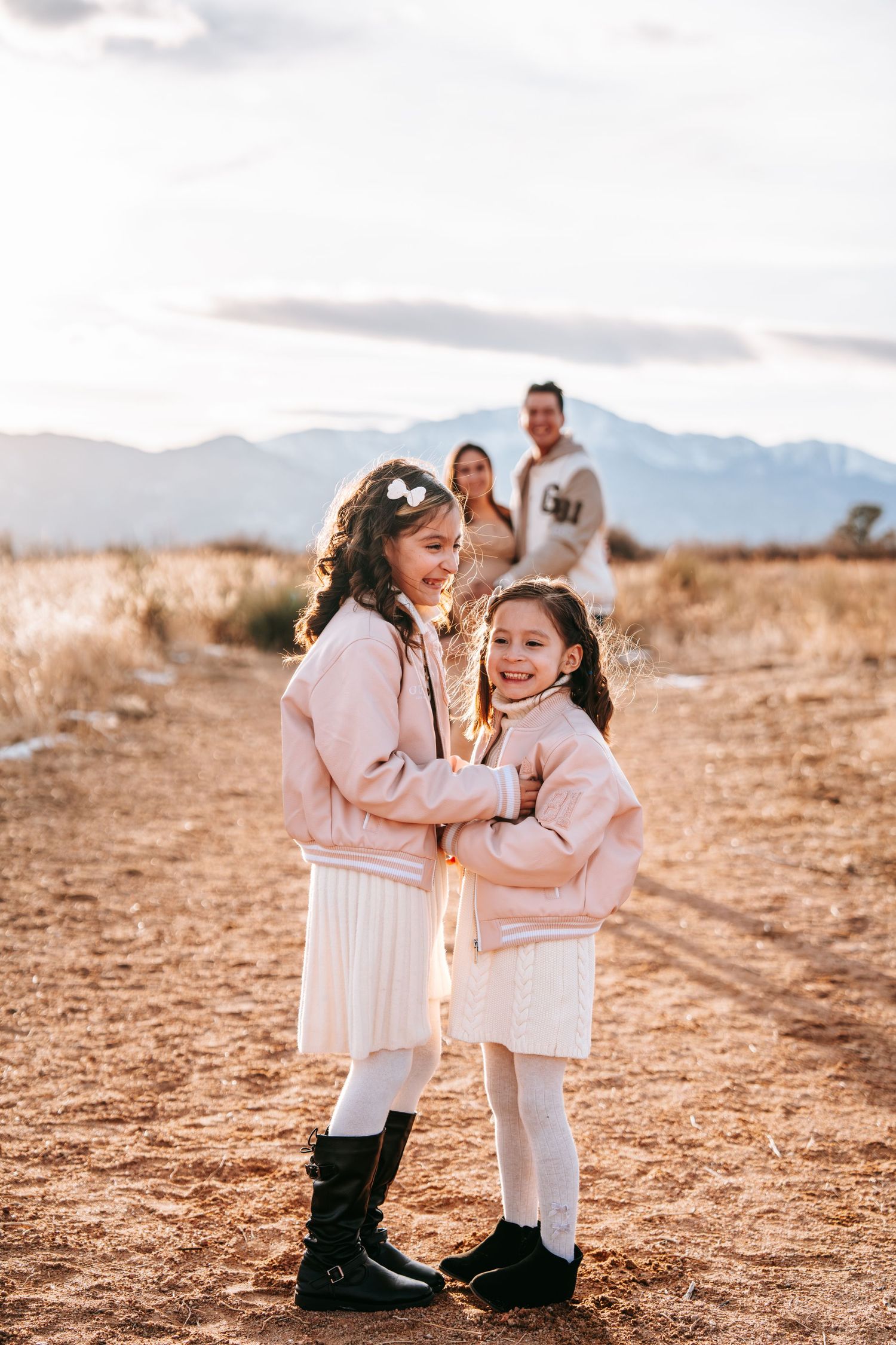 Two young sisters in matching pink sweaters smiling together outdoors with parents and mountains in the background.