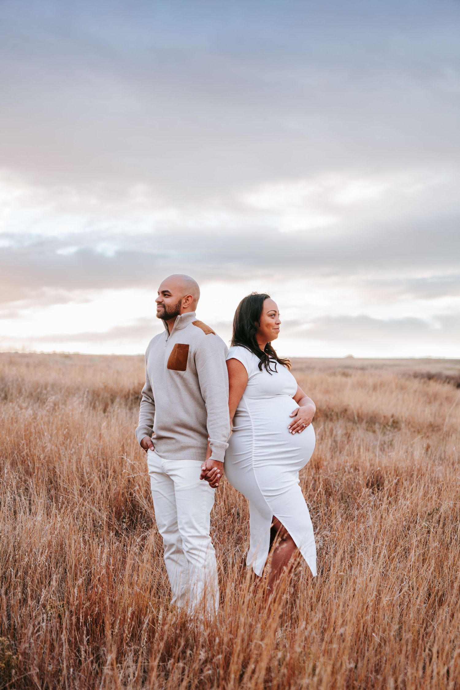 Expecting couple stands back to back holding hands in a golden grass field at sunset.