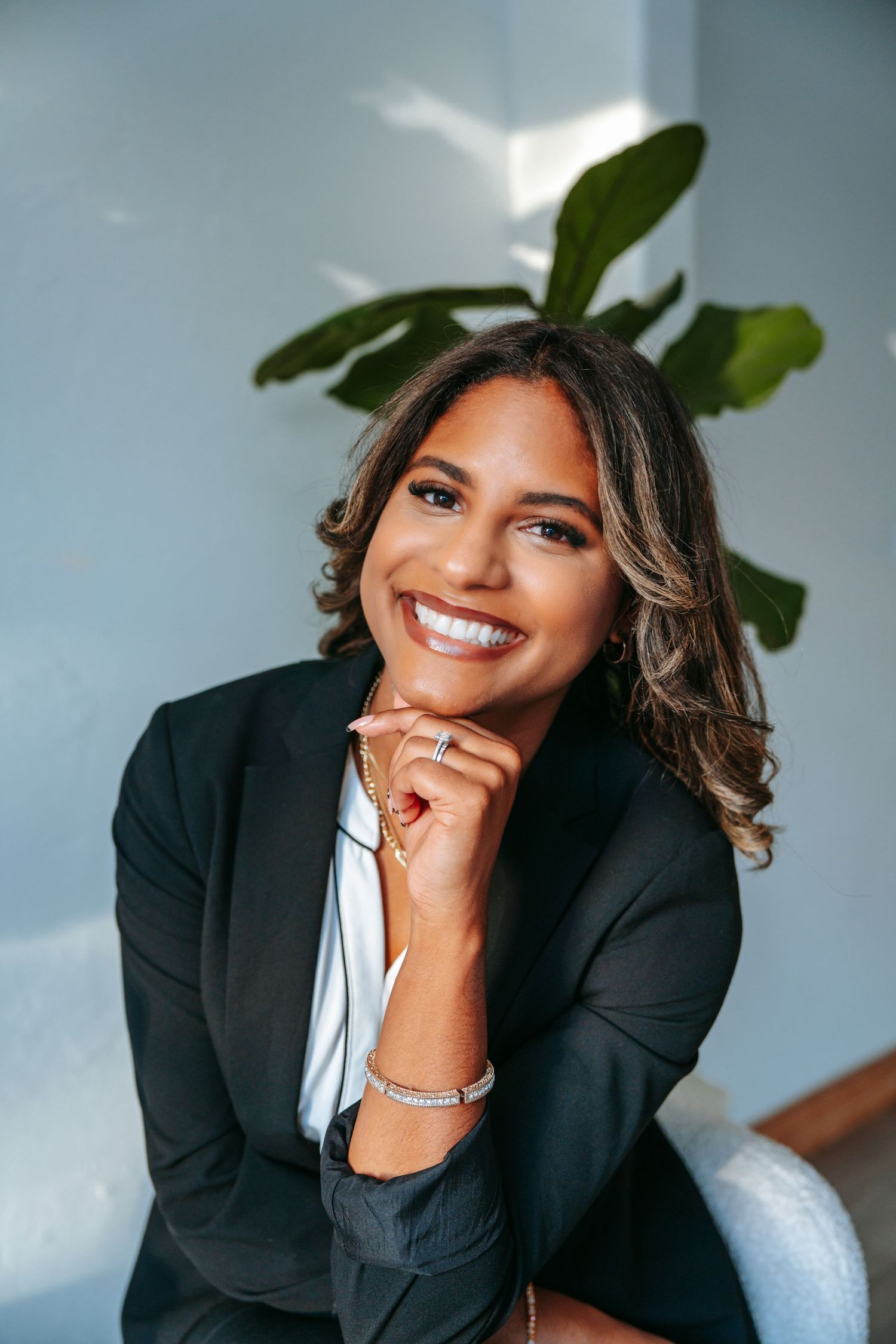 Smiling professional woman in black blazer posing with hand on chin beside a large green fiddle leaf fig plant.