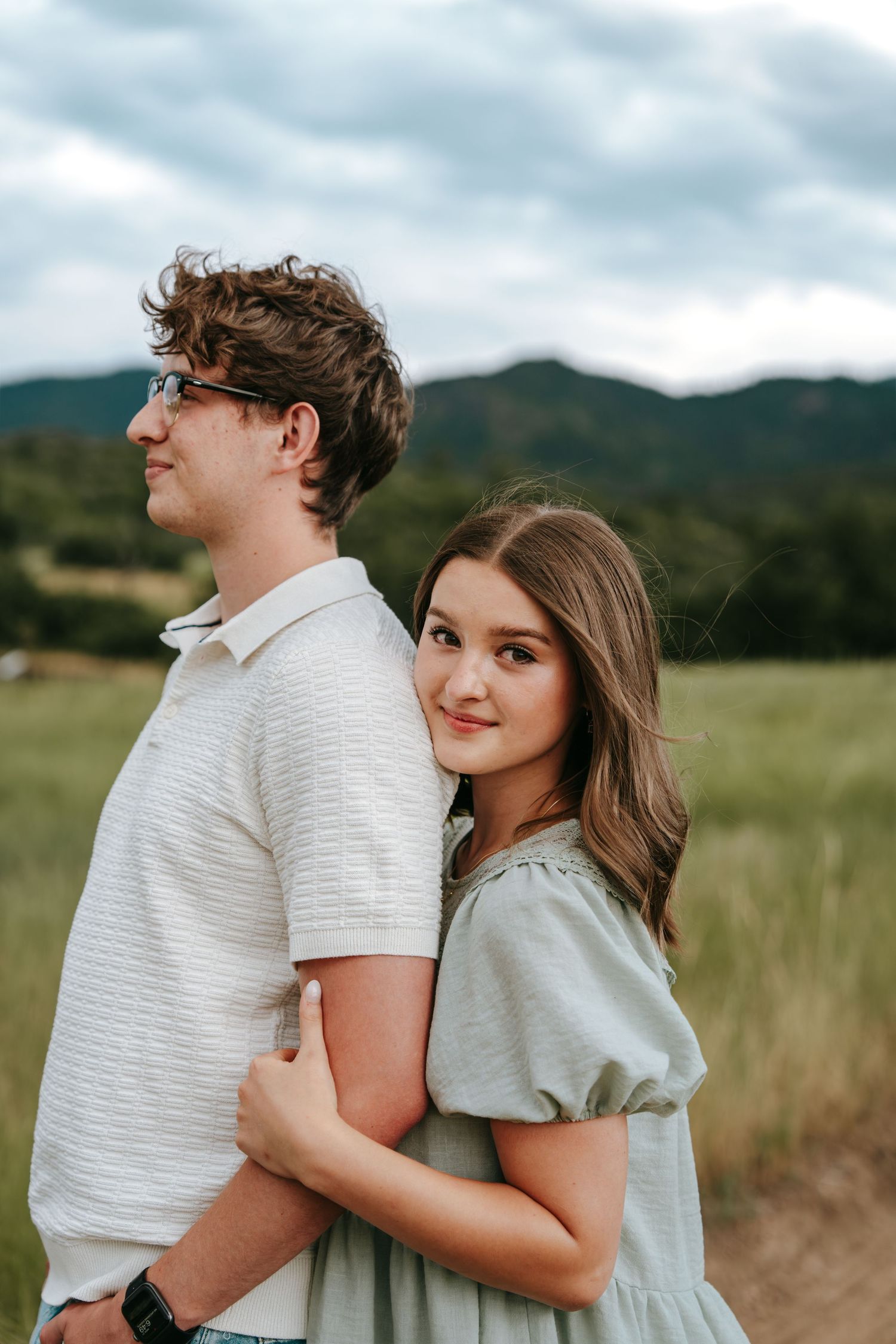 Young woman hugging man from behind in a mountain meadow, smiling softly at camera.