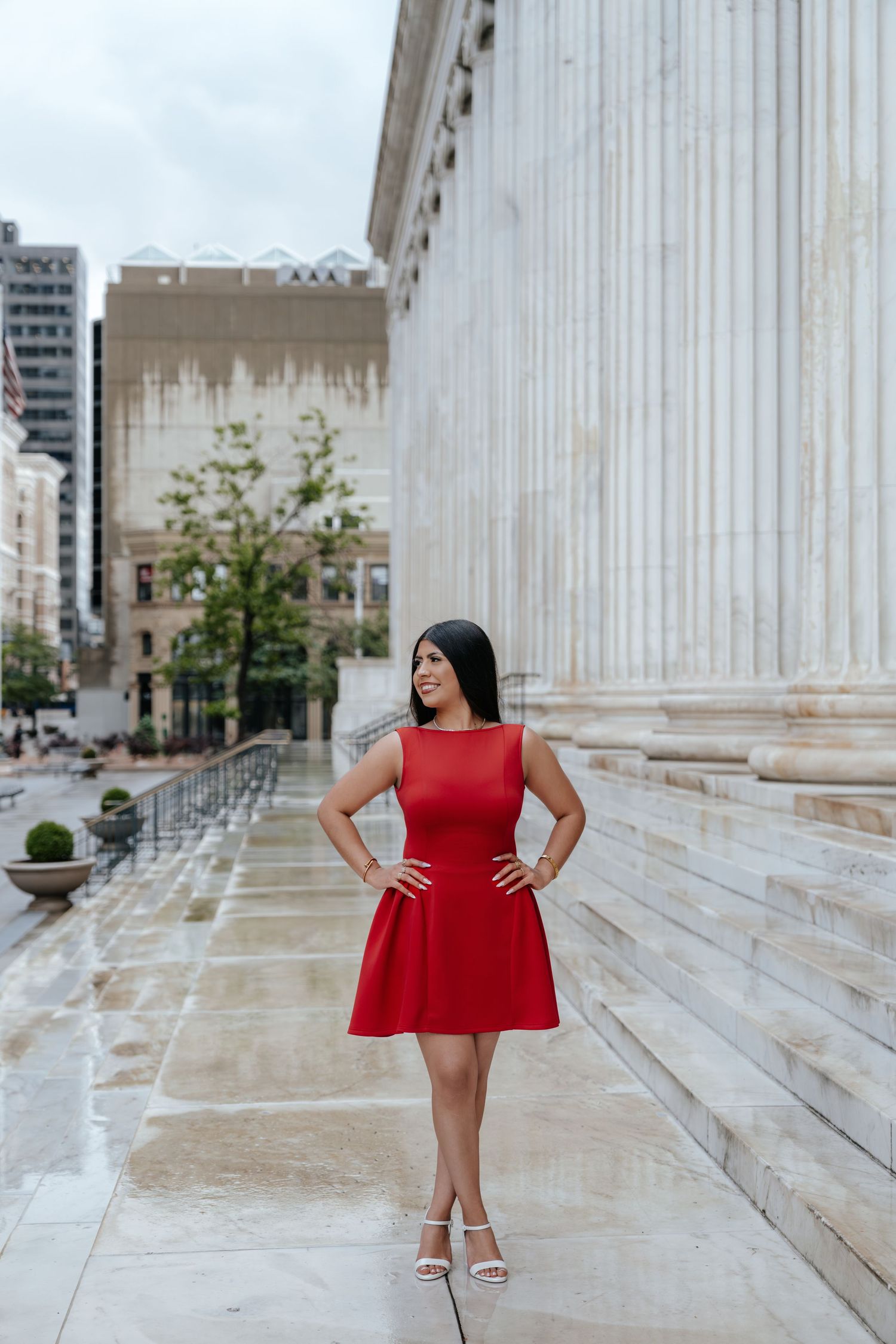 Young woman in red dress poses confidently with hands on hips beside a large marble column on stone steps.